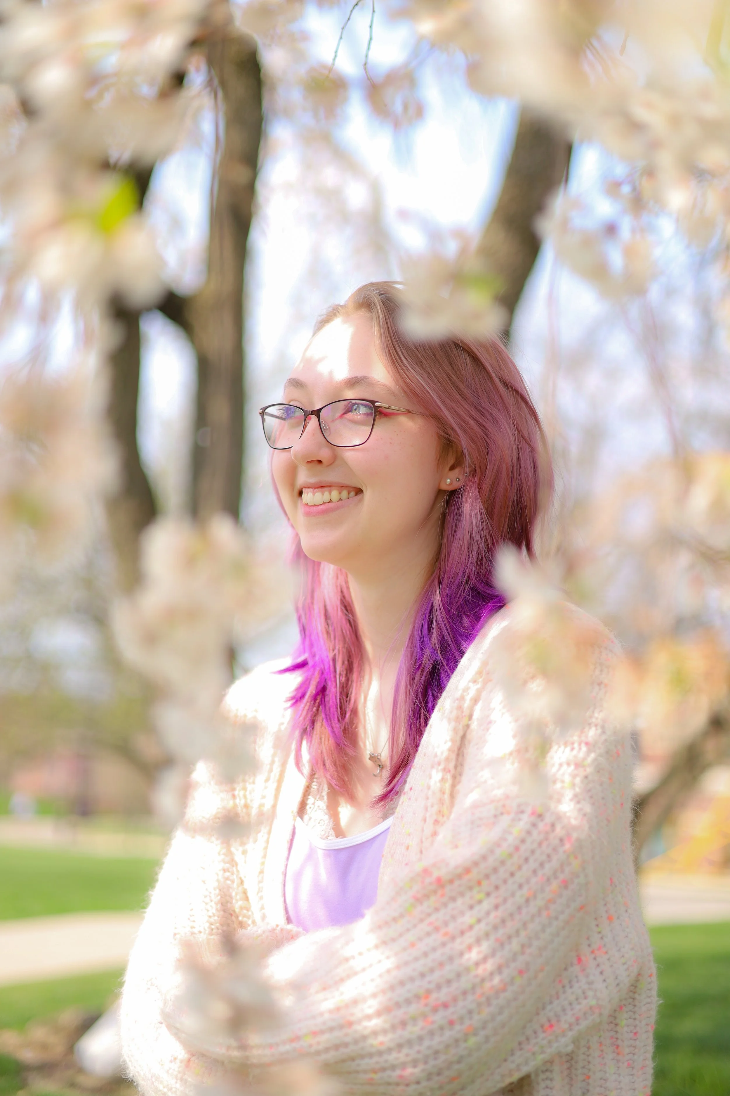 Smiling young woman with glasses and pink-purple hair standing among blooming tree branches on a sunny day.