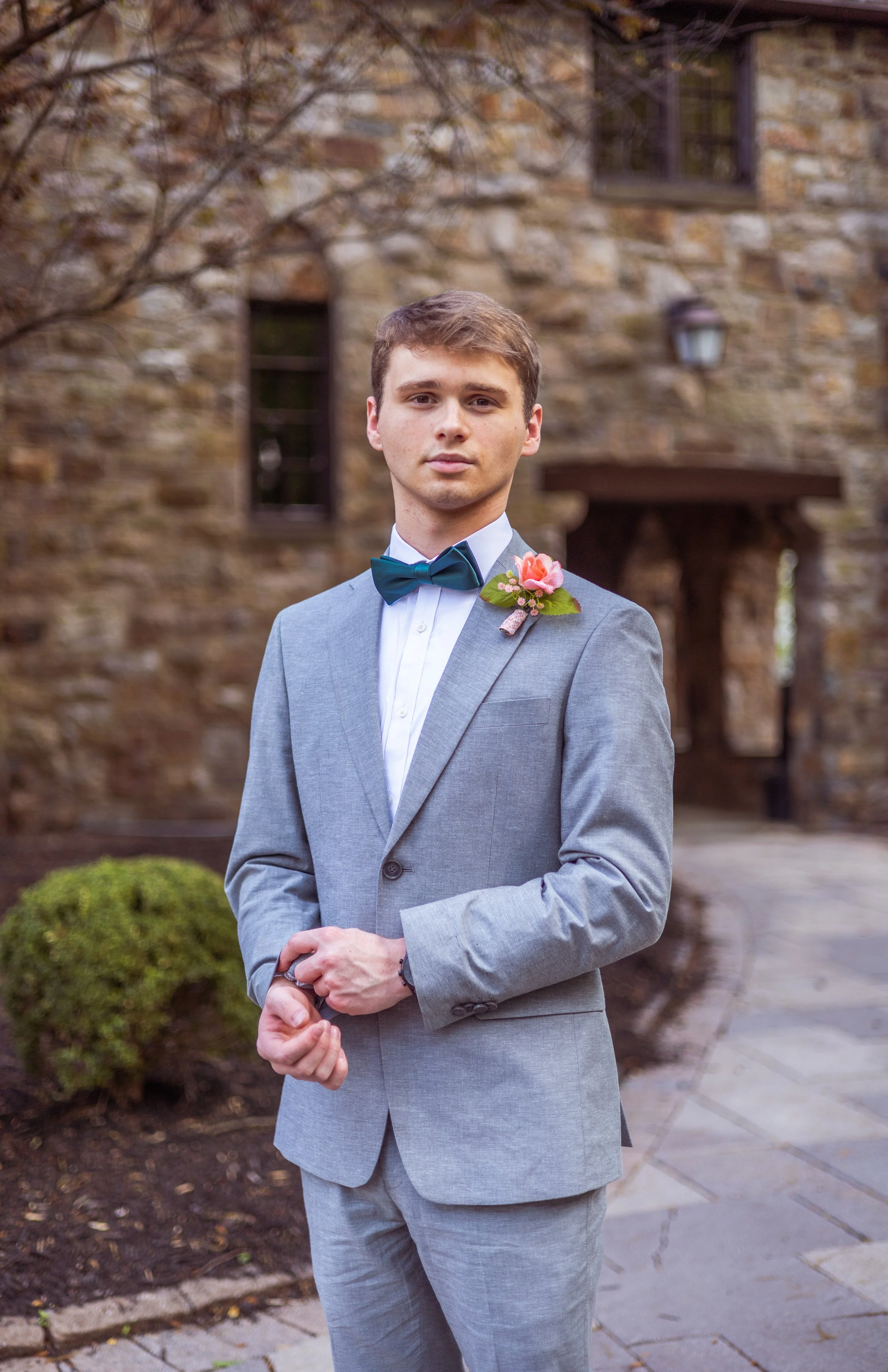 Young man in a gray suit with a teal bow tie, standing outdoors in front of a stone building, wearing a pink flower boutonniere. He has short brown hair and a serious expression.