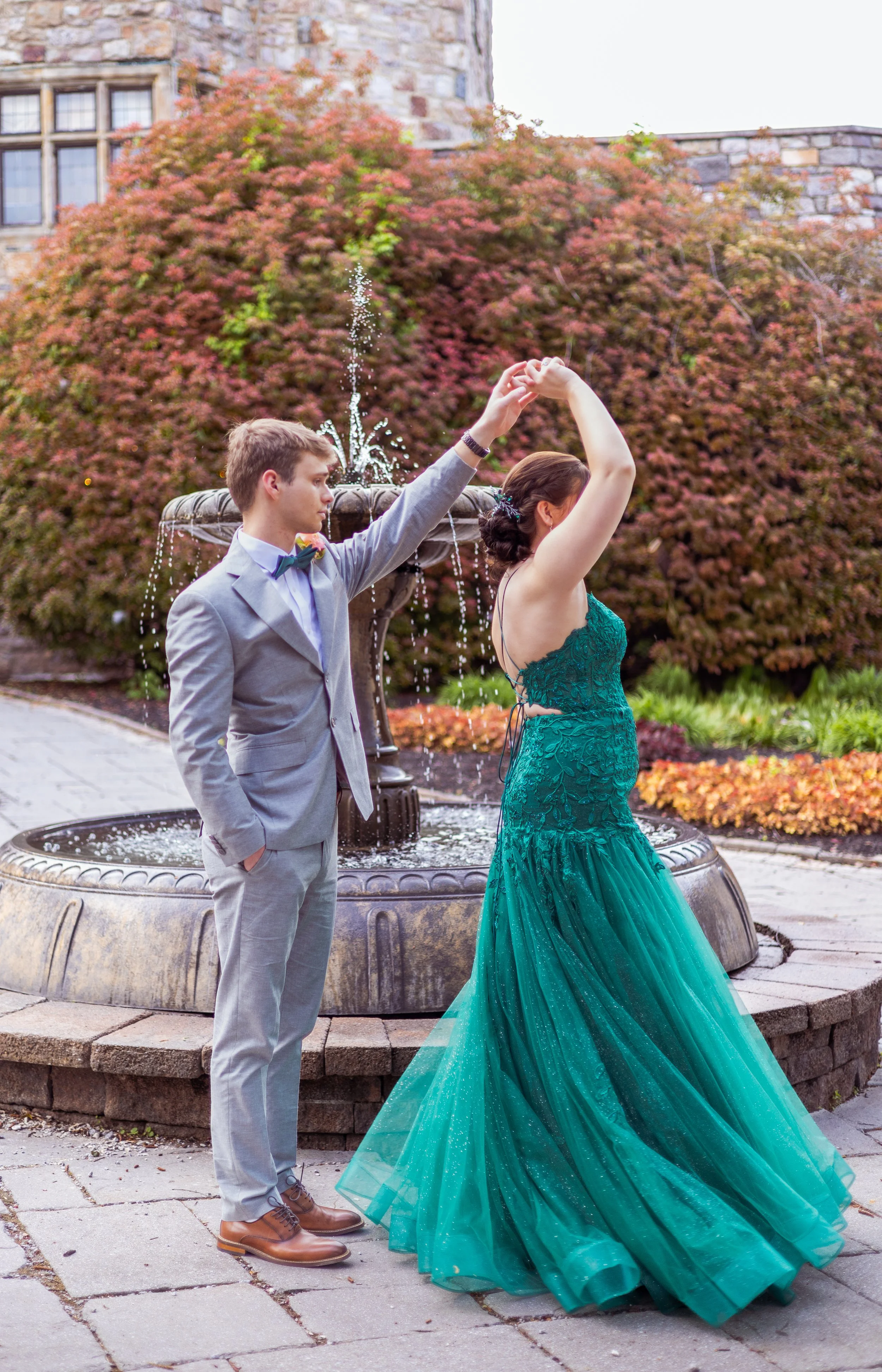A young couple dancing by a fountain in an outdoor garden during autumn, with the man in a light gray suit and the woman in a teal gown.