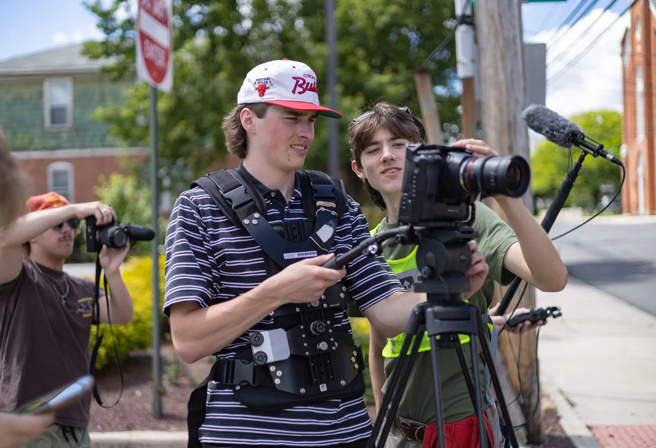 Two young men, one wearing a Chicago Bulls hat, operate a professional camera on a tripod while filming outdoors, with a third person partially visible recording with a handheld camera nearby.