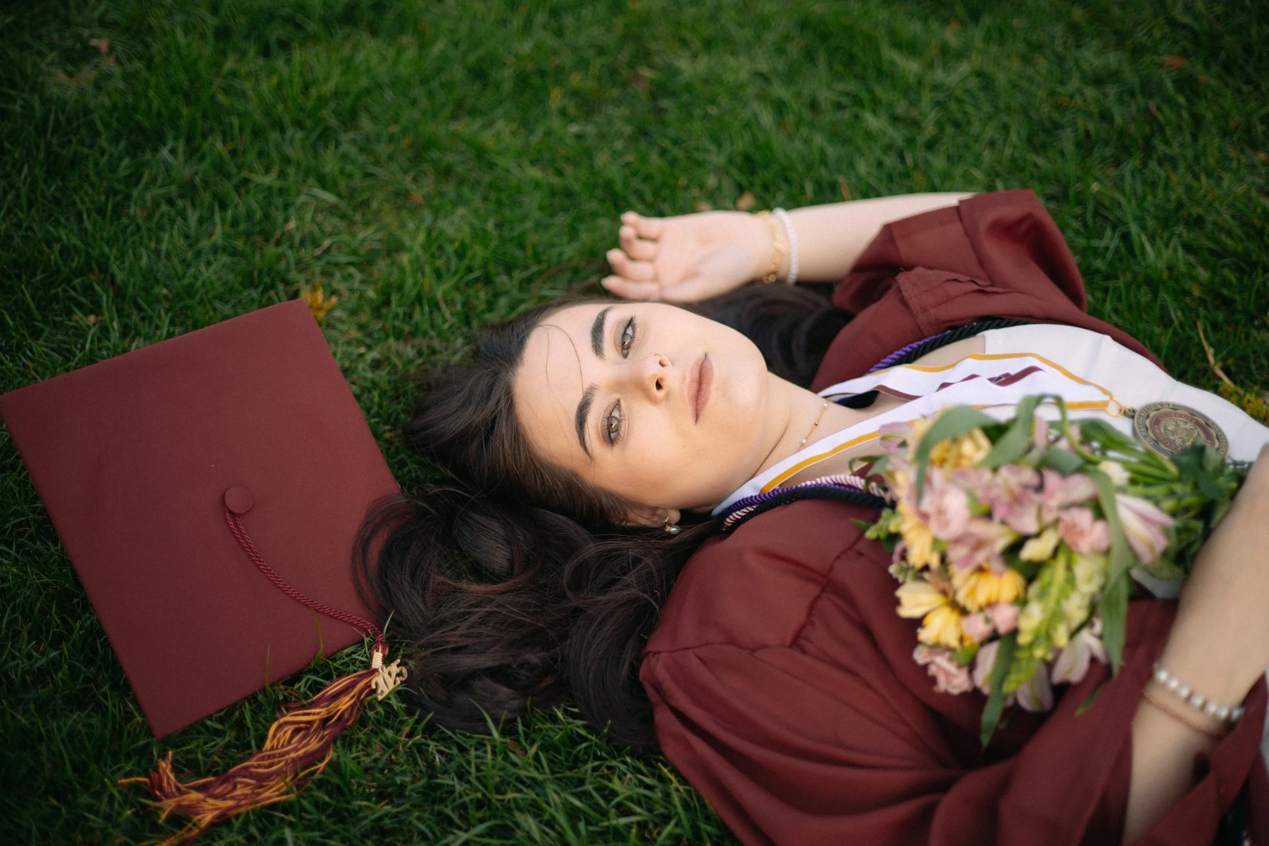 A young woman lying on green grass, wearing a graduation gown and cap, holding a bouquet of flowers, with her graduation medal visible, and her graduation cap beside her.