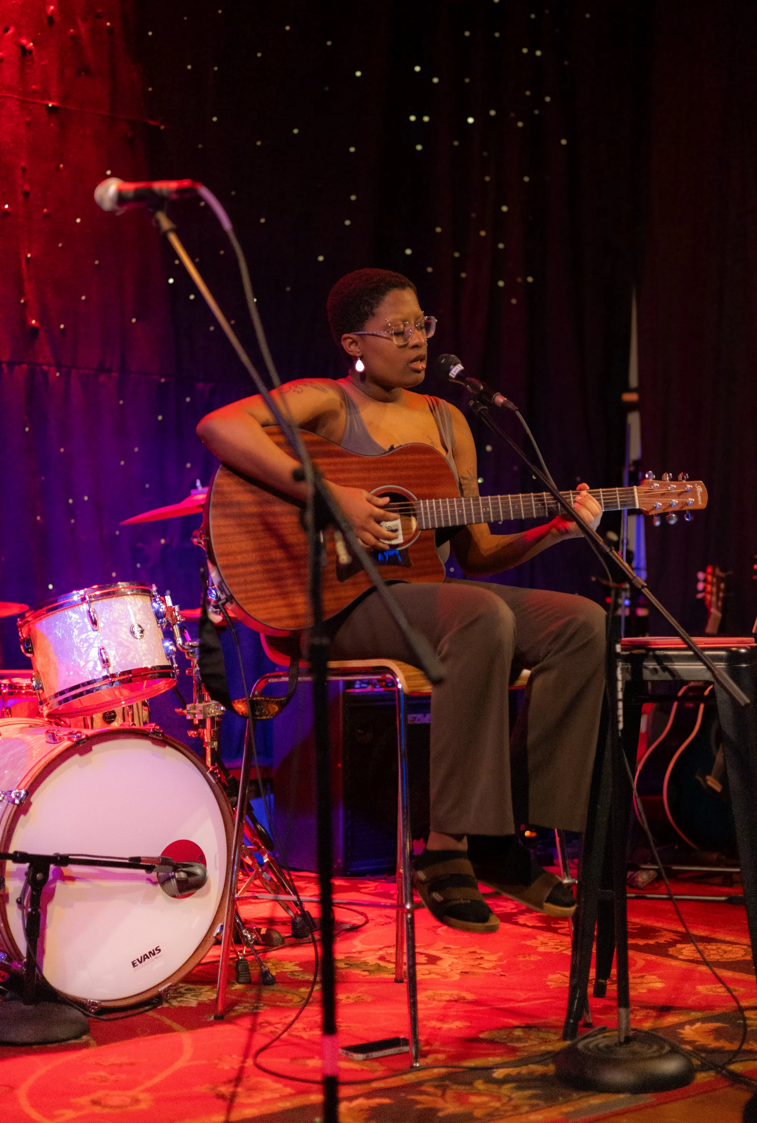 A woman with glasses and tattoos playing an acoustic guitar on stage, with a drum set behind her, during a musical performance.