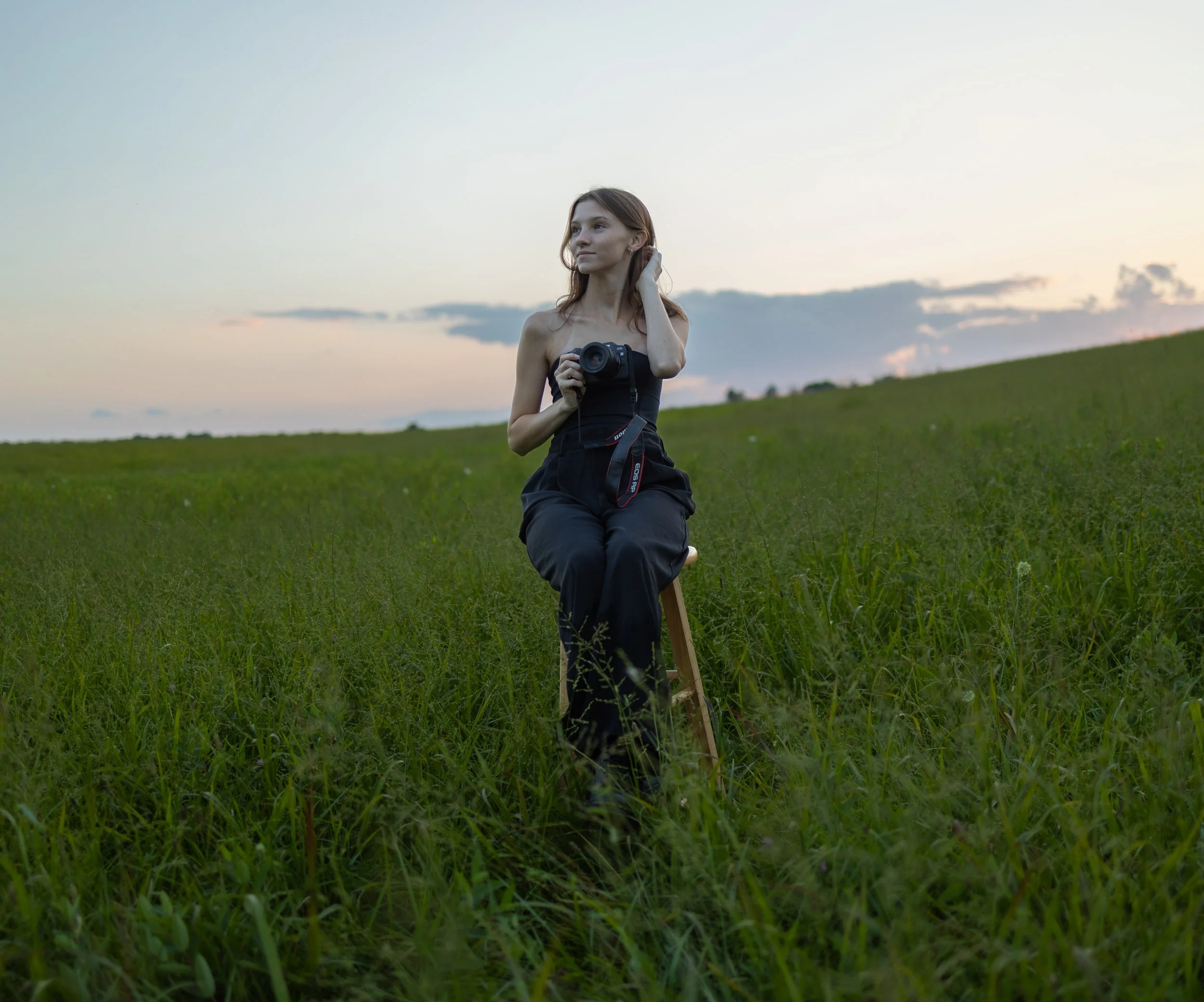 A woman in a black outfit sitting on a wooden stool in a grassy field during sunset, holding a camera and touching her hair.