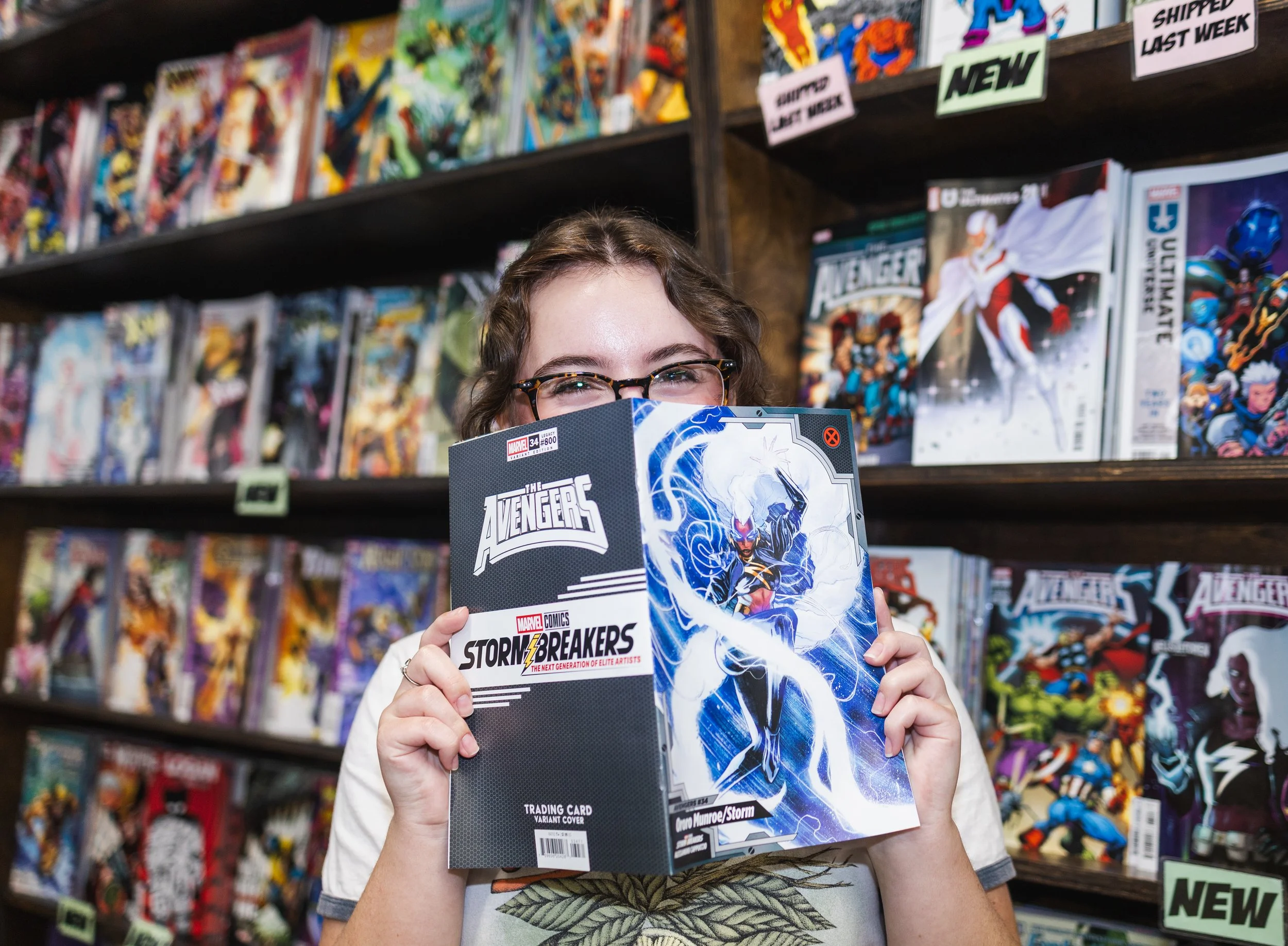 A young woman with glasses holding a Marvel comic book titled "Stormbreakers" in front of a comic book shelf in a store.