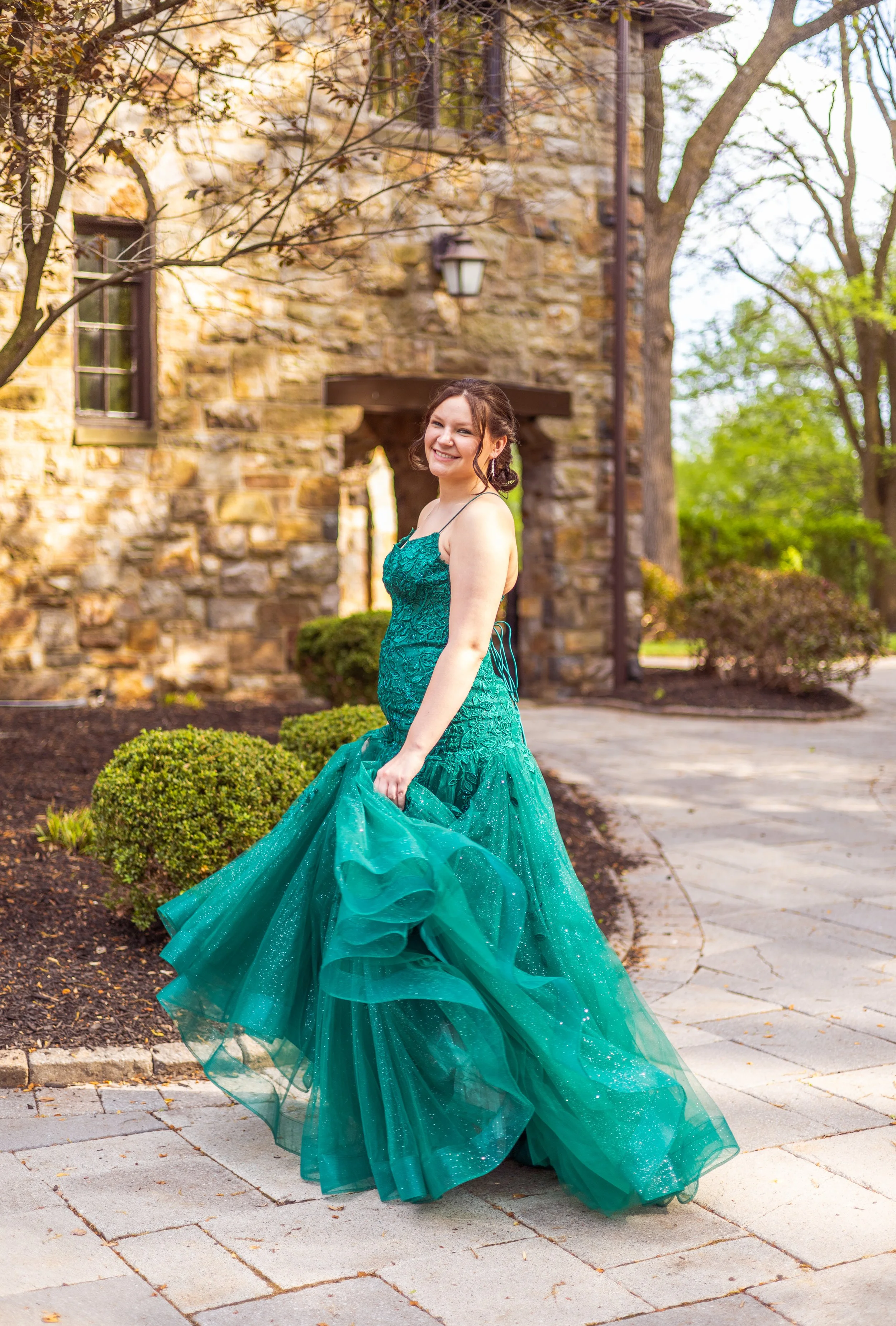 A young woman in a teal gown standing outdoors on a paved walkway in front of an old stone building with trees and bushes surrounding her, smiling at the camera.