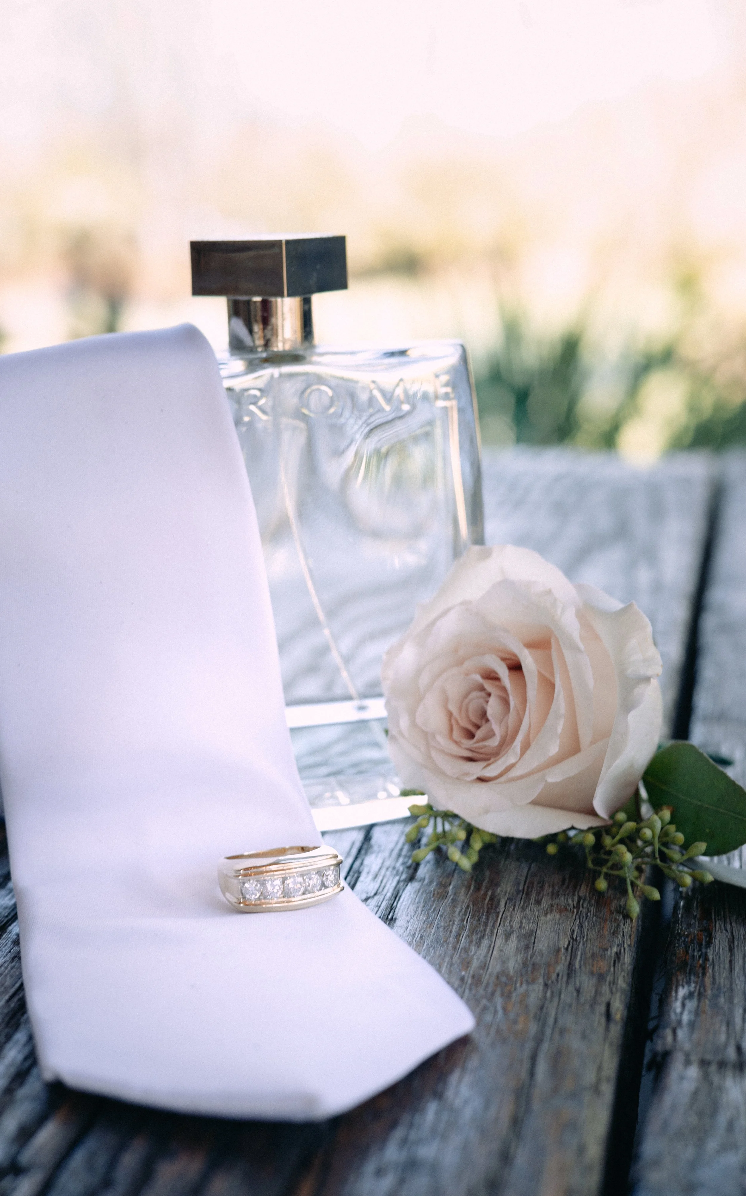 A wedding ring on a white napkin, a single cream-colored rose, a glass perfume bottle, and a blurred outdoor background.