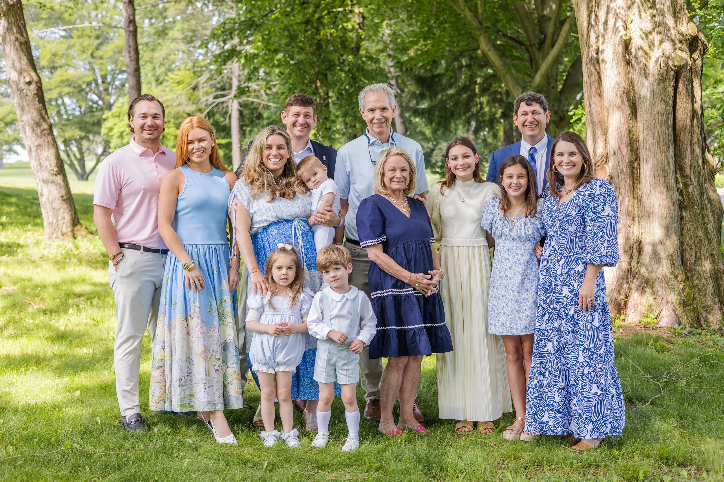 A large multigenerational family posing outdoors under trees on a sunny day, dressed in pastel and summer outfits.