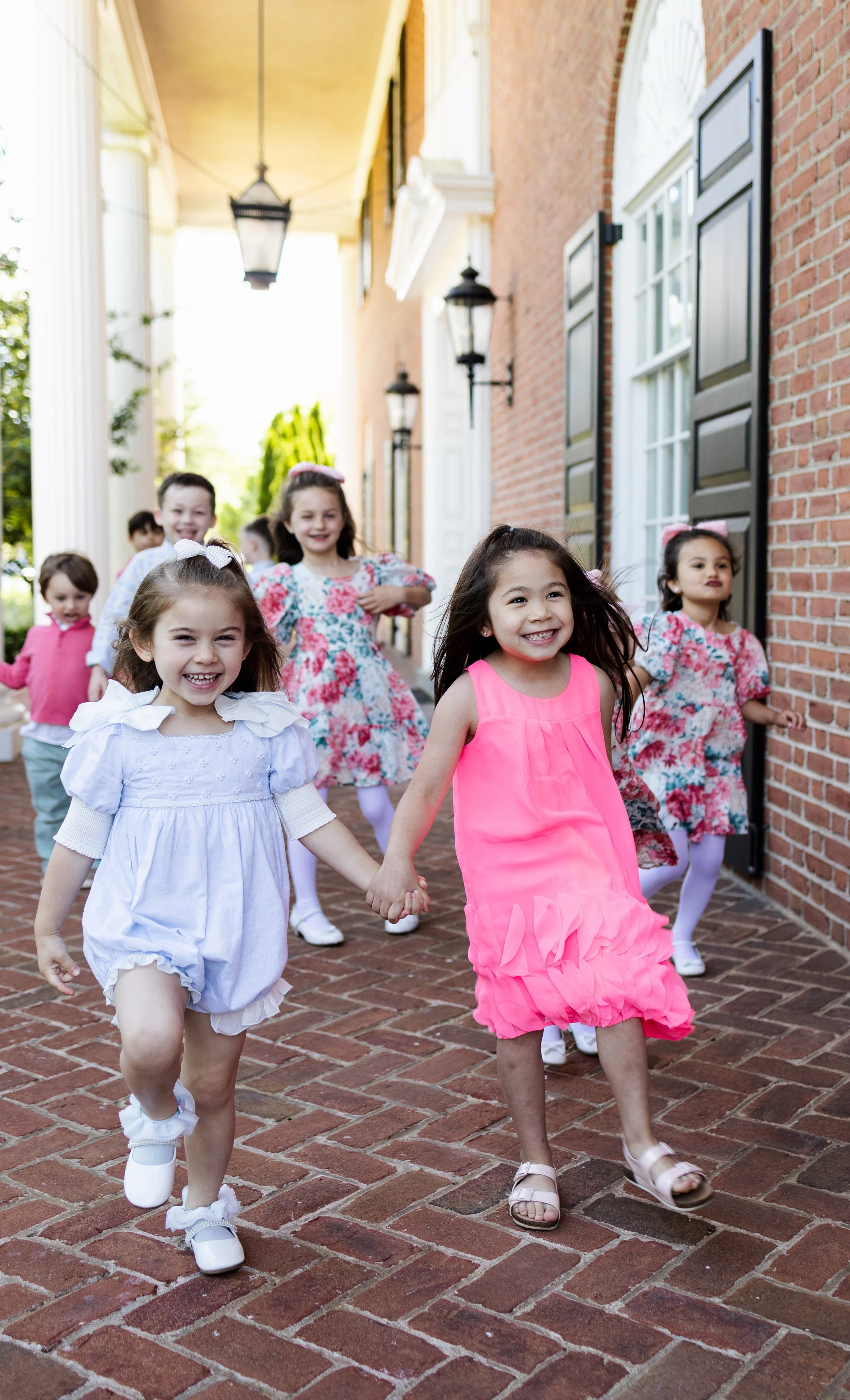 Group of young children in colorful dresses happily running outside on brick pavement.