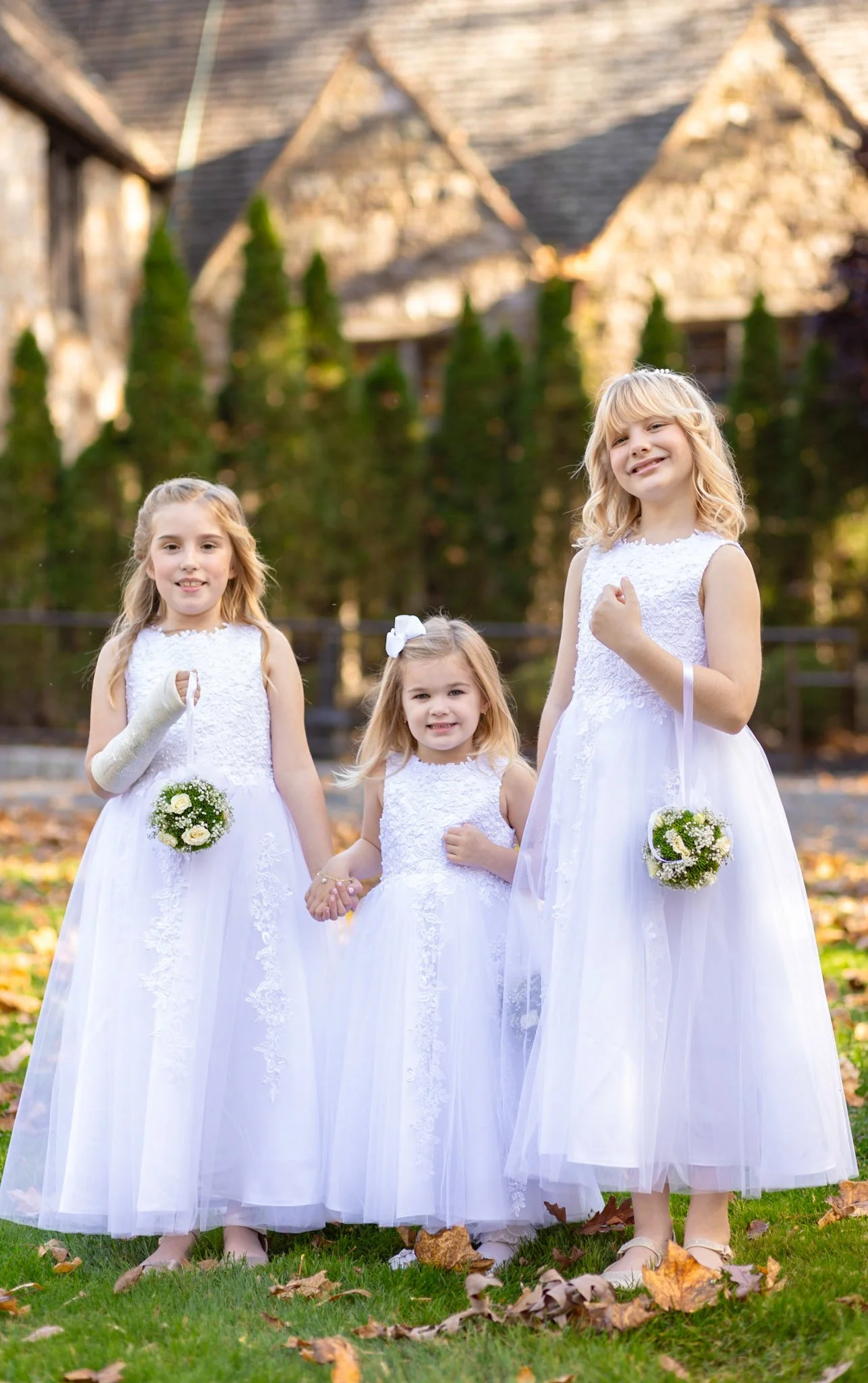 Three young girls in white dresses holding flower bouquets outdoors on grass with fallen leaves during daytime.