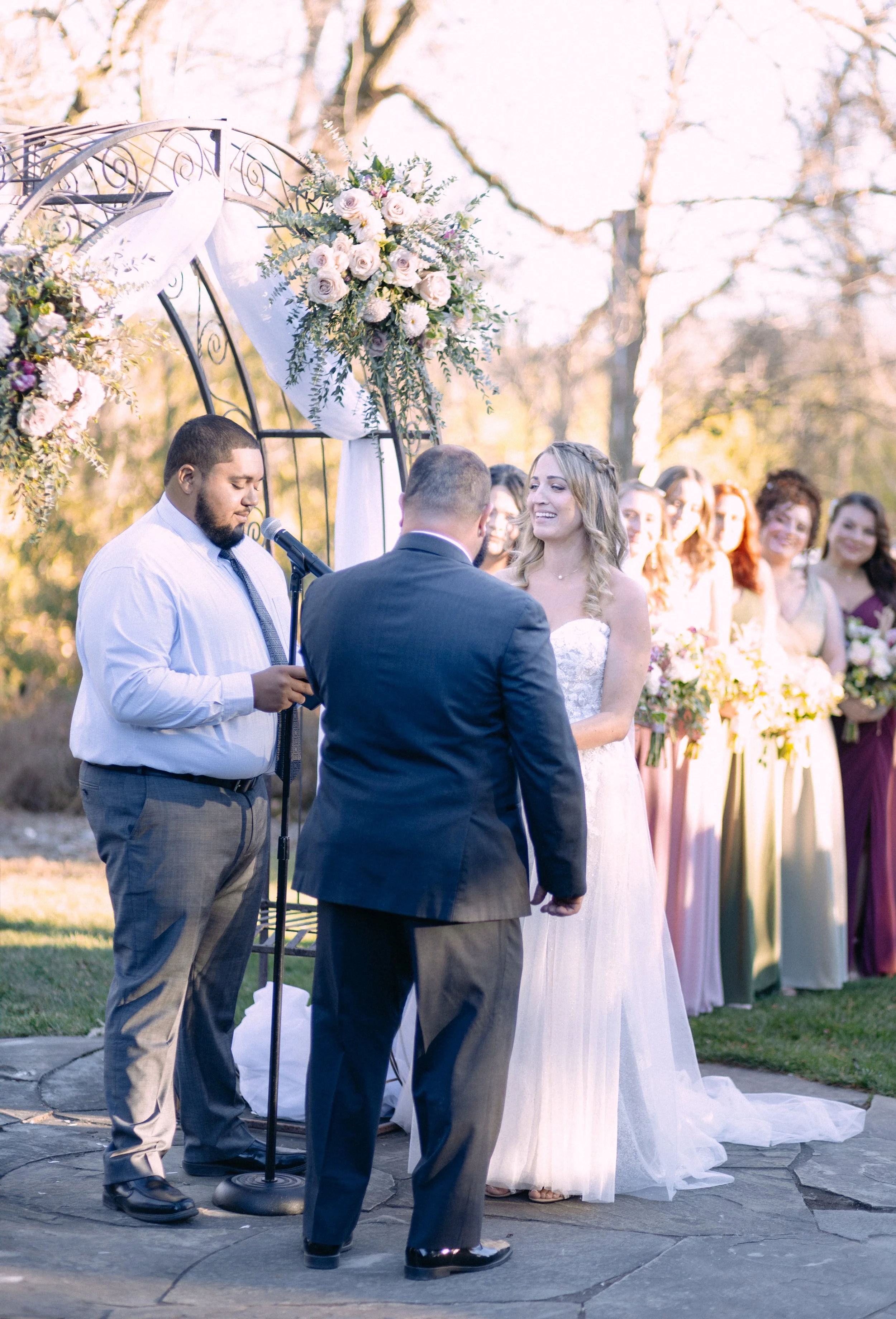 A couple making wedding vows during an outdoor wedding ceremony, with a officiant and bridesmaids in pastel dresses holding bouquets