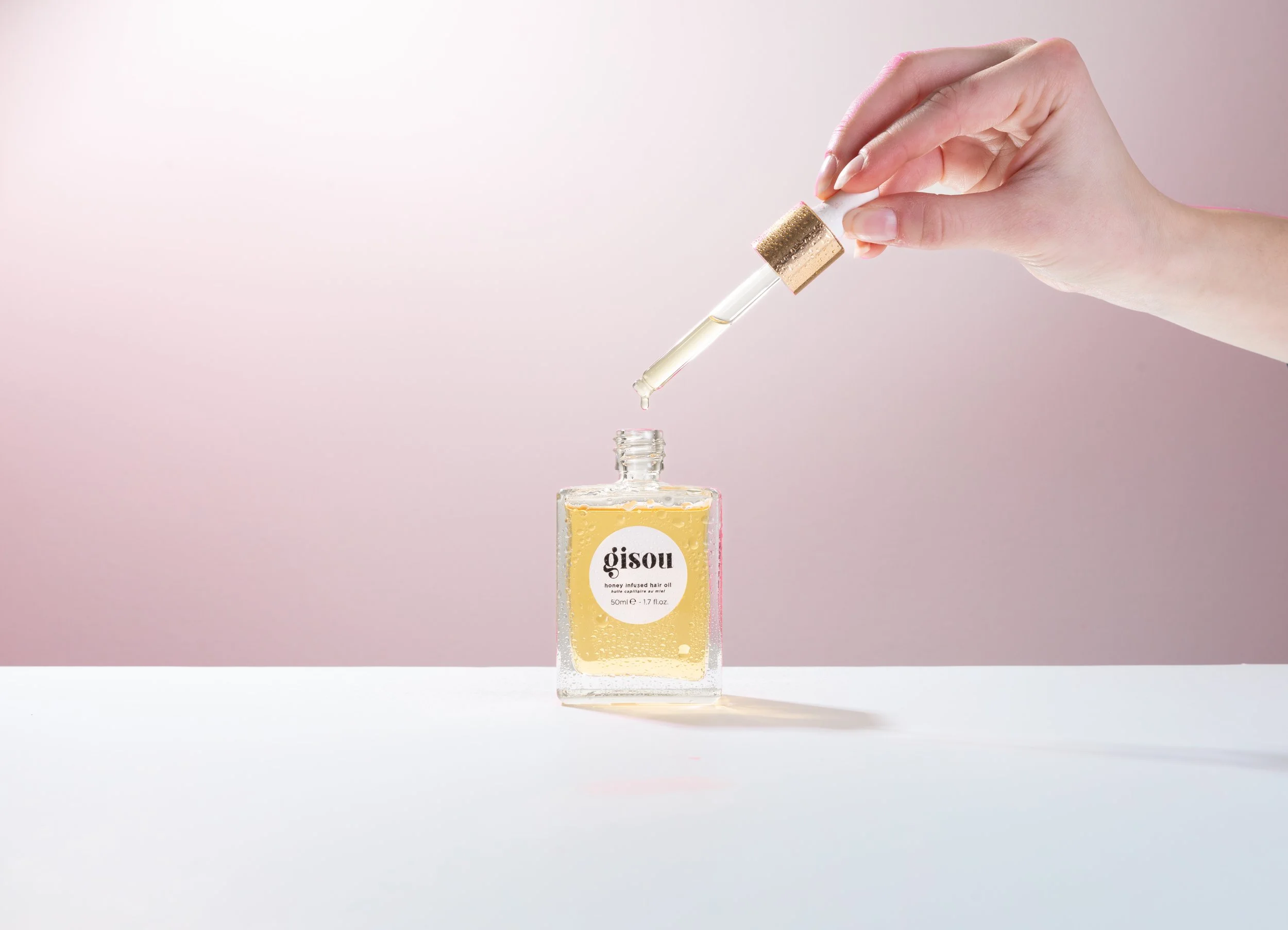 A hand holding a dropper above a glass bottle filled with a yellow liquid, labeled 'Gisou honey infused hair oil,' on a white surface with a pink background.