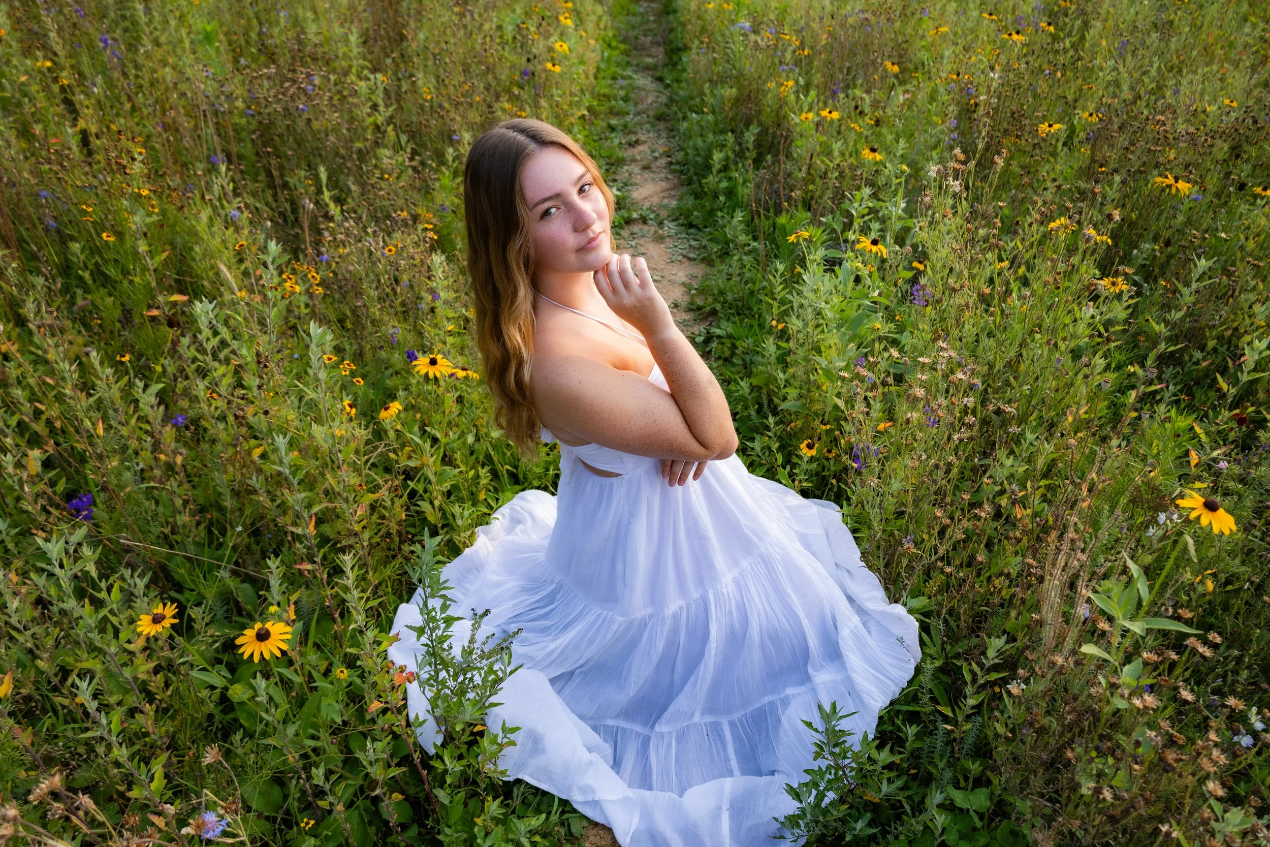 A young woman in a white dress sitting on a dirt trail amidst wildflowers in a green field.