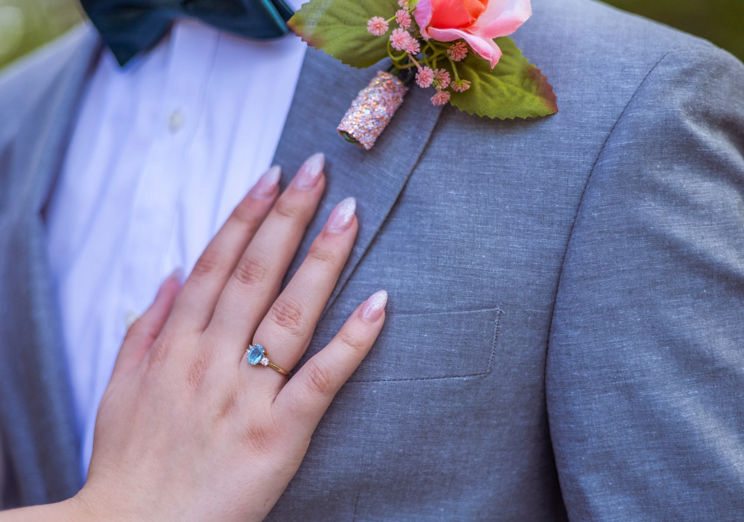Close-up of a woman's left hand with an engagement ring featuring a blue gemstone, resting on a man's gray suit jacket. The man is wearing a white shirt and a dark bow tie, with a floral boutonniere featuring pink flowers and green leaves pinned to h