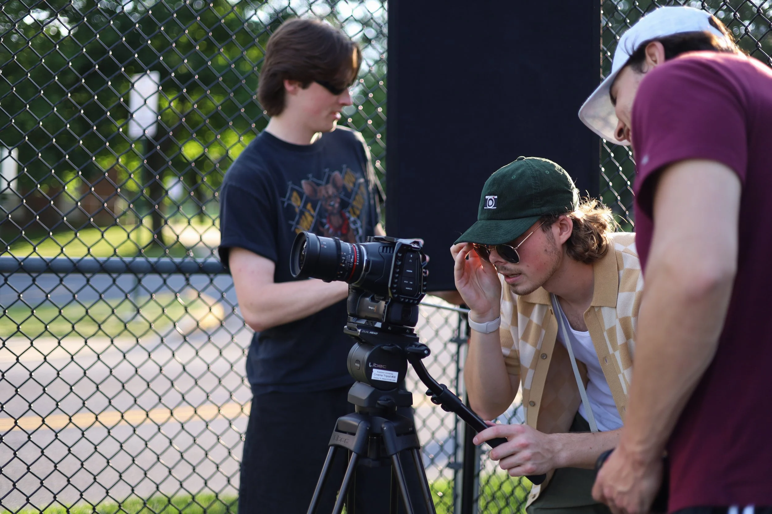 Three young men, one with sunglasses and a green cap, operate a camera on a tripod behind a chain-link fence, with another person in glasses and a salmon-colored shirt leaning in to observe, outdoors during daytime.