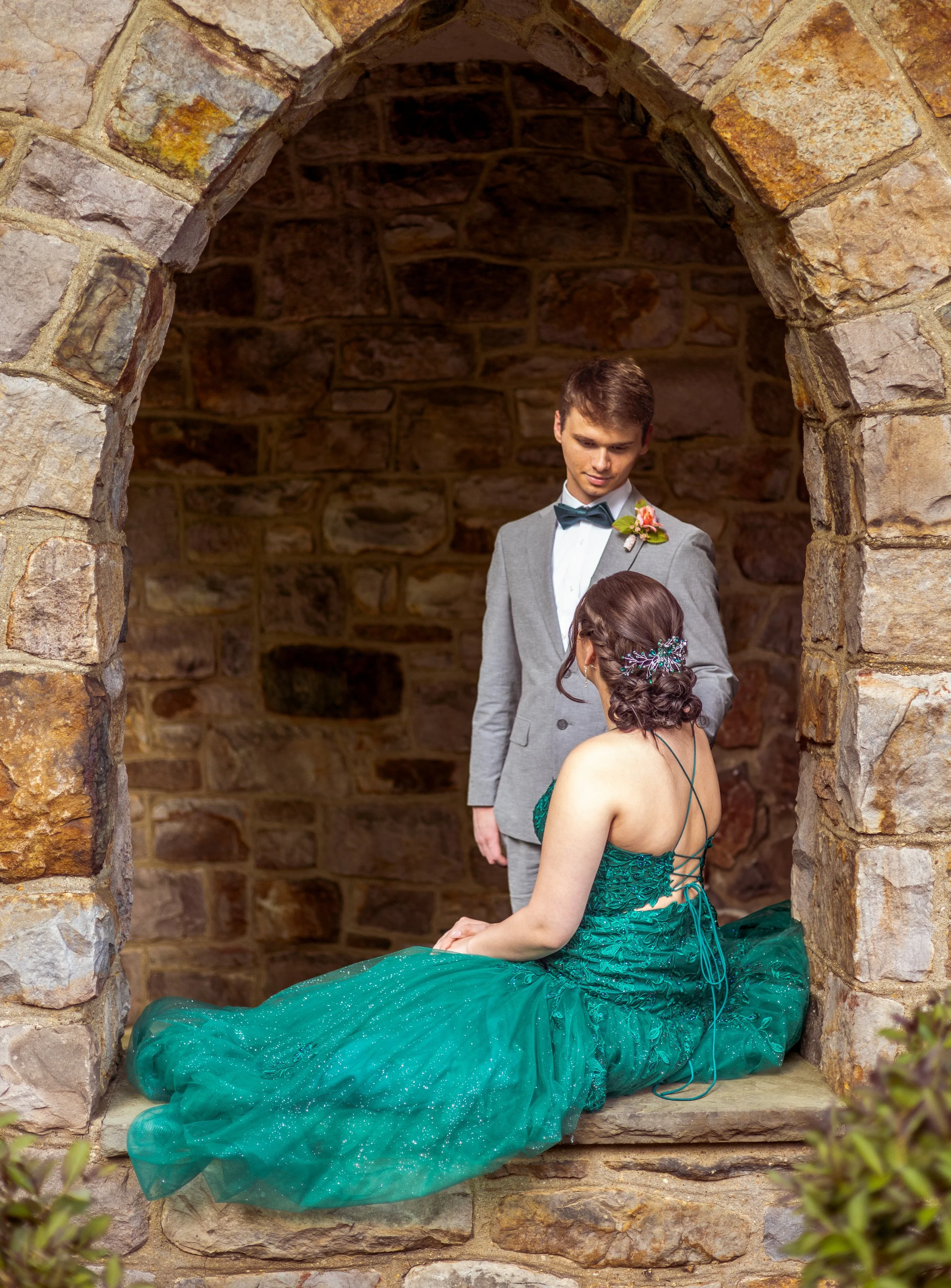 A man and woman in formal attire inside a stone alcove. The woman is seated wearing a green gown with intricate details, and the man is standing in a light gray suit with a bow tie, looking at her.
