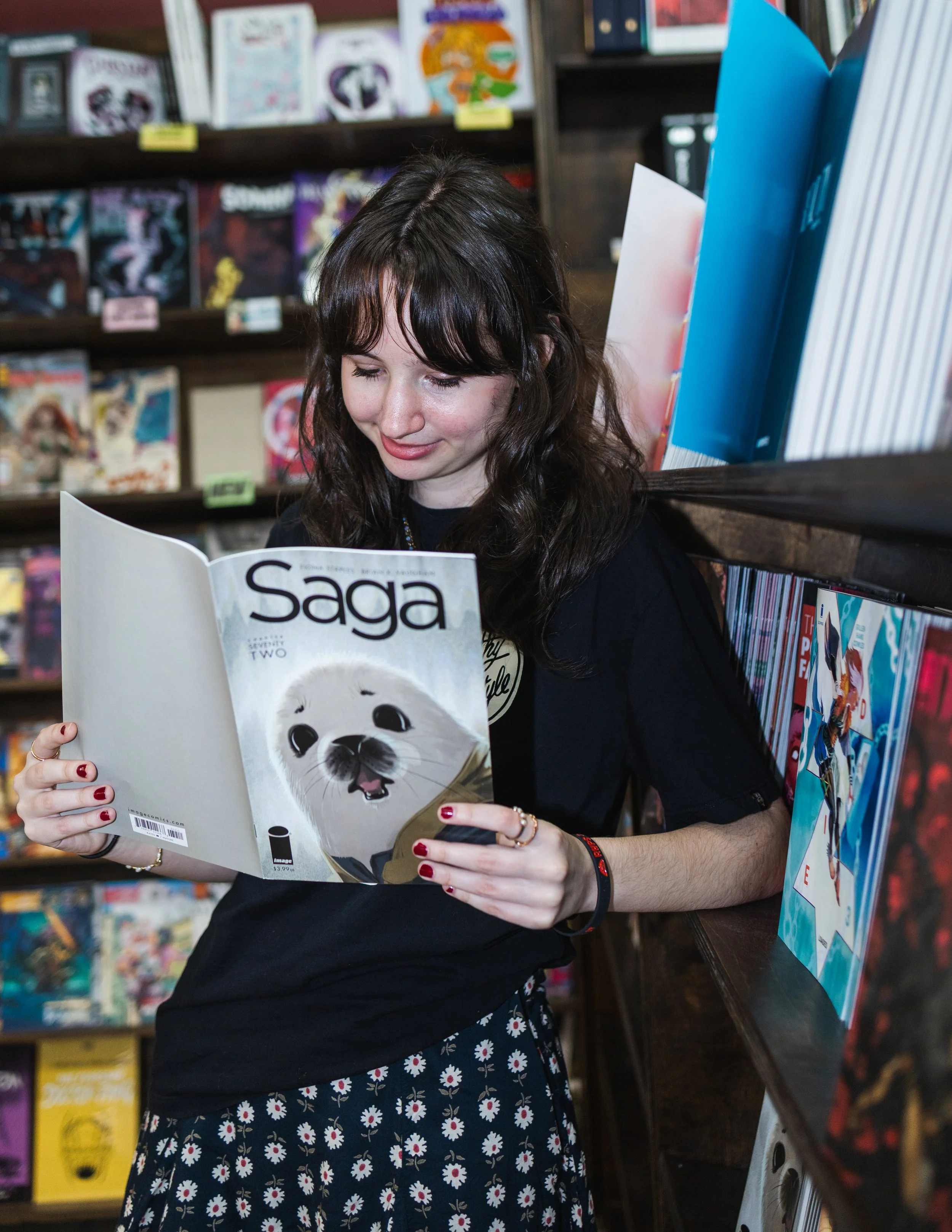 A young woman with dark, wavy hair and a black shirt is reading a comic book called 'Saga' with a white dog on the cover at a bookstore, surrounded by shelves of comics.