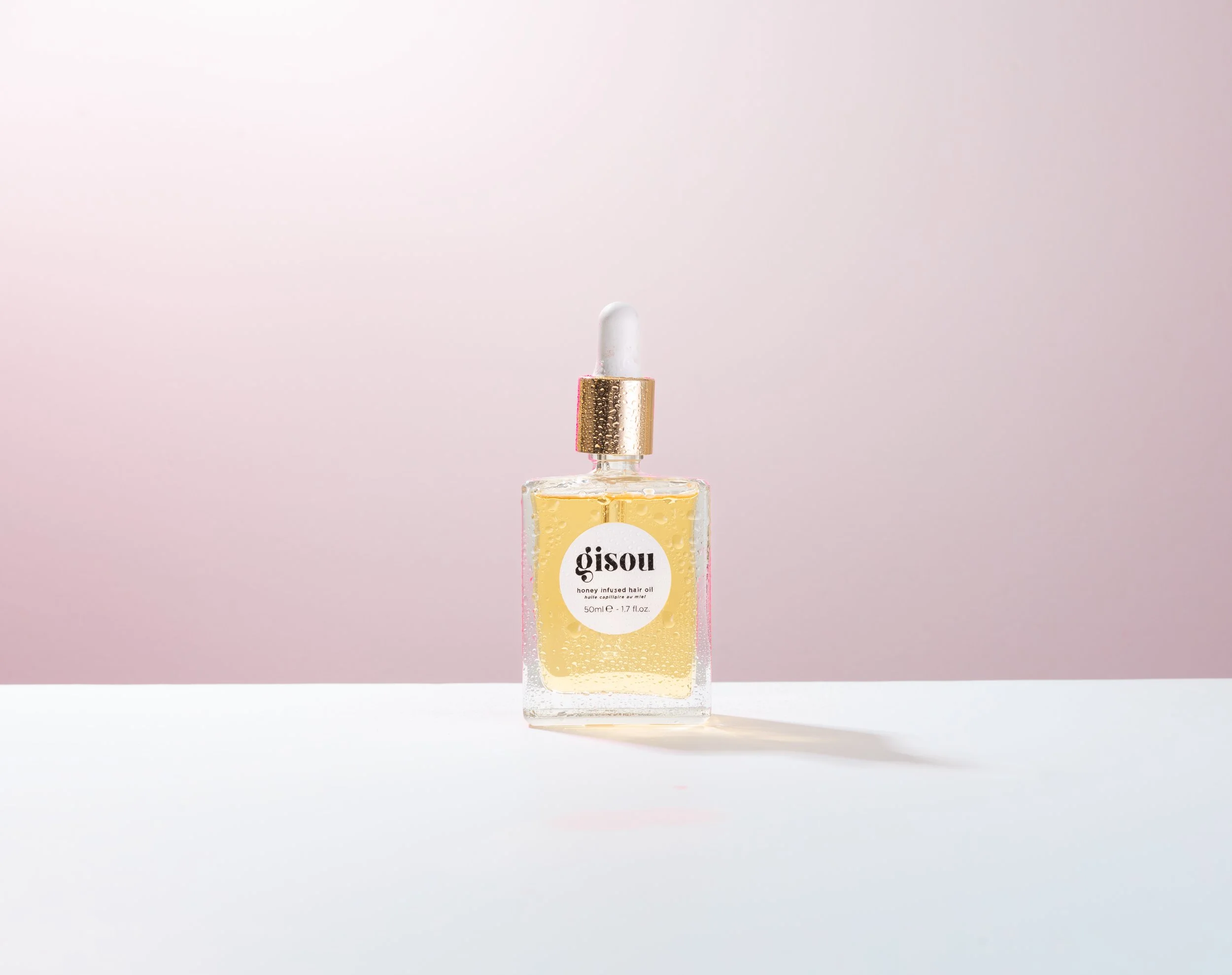 Bottle of Gisou honey infused hair oil with a gold dropper cap on a white surface against a pink background.
