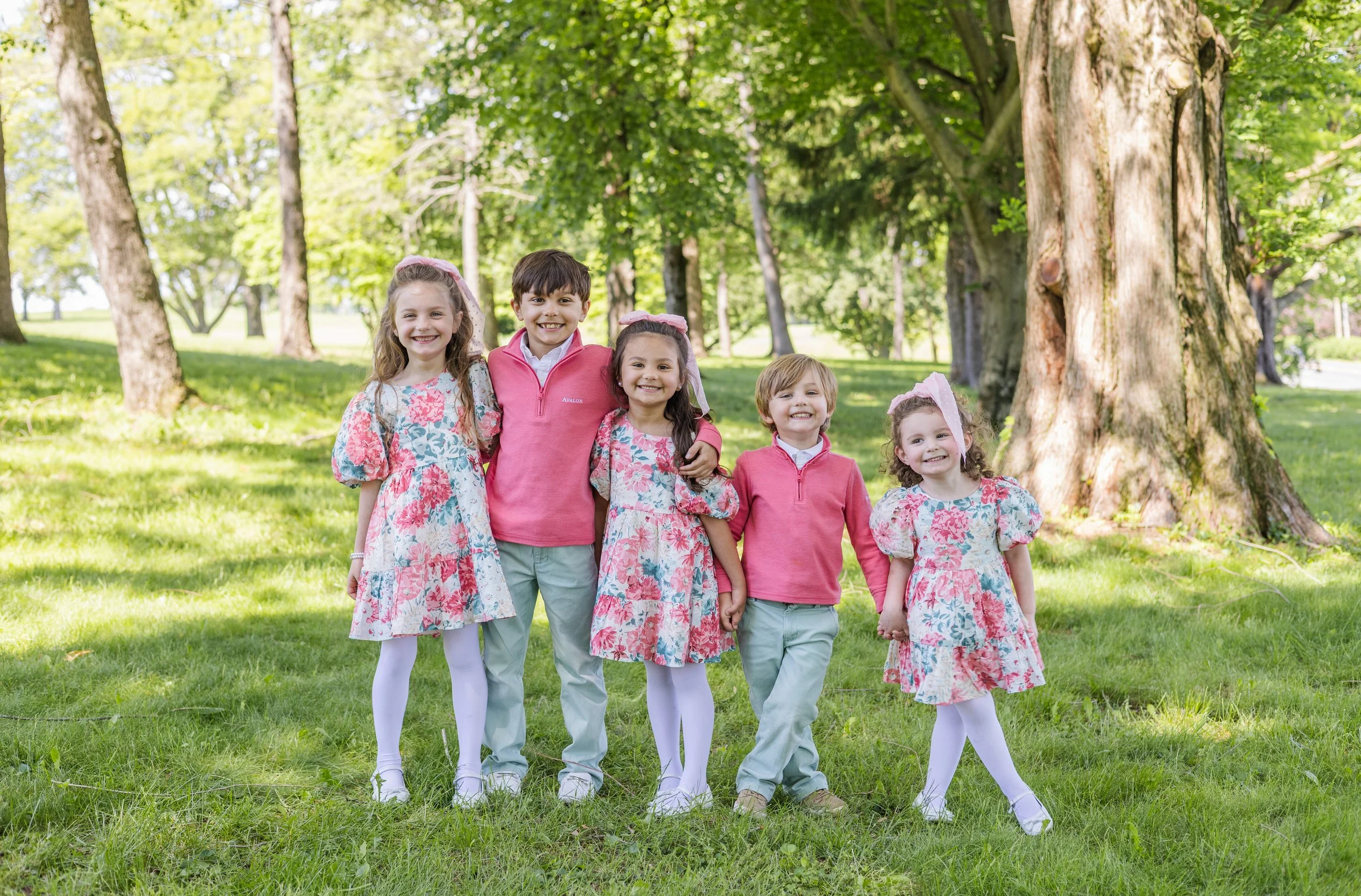 Five children standing outdoors in a park with green trees and grass, smiling and holding hands.