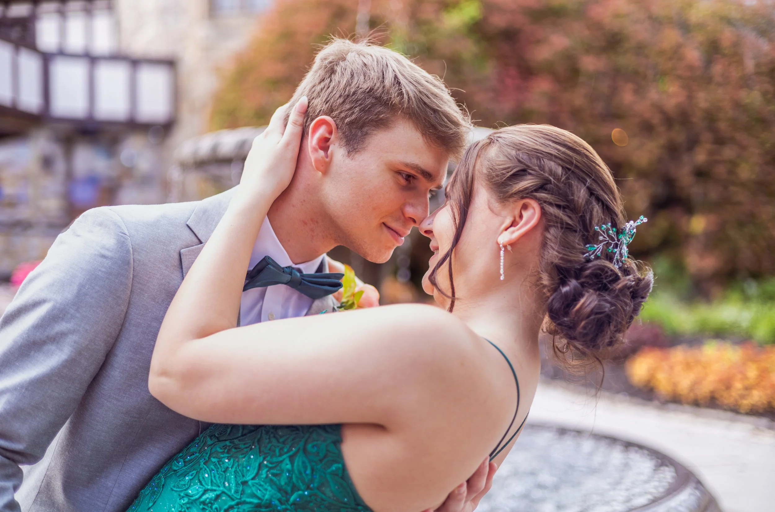 A young couple dressed in formal attire, sharing an intimate moment outdoors with their foreheads touching, surrounded by autumn foliage.