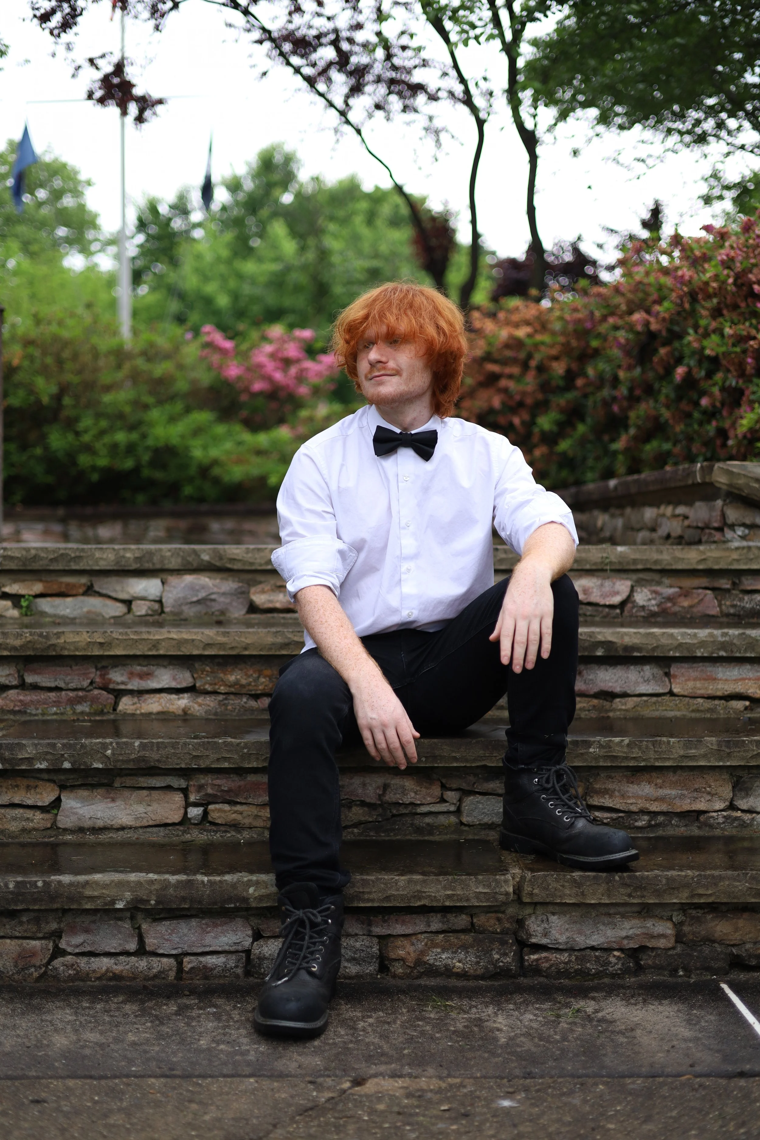 A young man with curly red hair and freckles, wearing a white shirt with rolled-up sleeves, a black bow tie, black pants, and black boots, sitting on stone steps outdoors surrounded by greenery and flowering bushes.