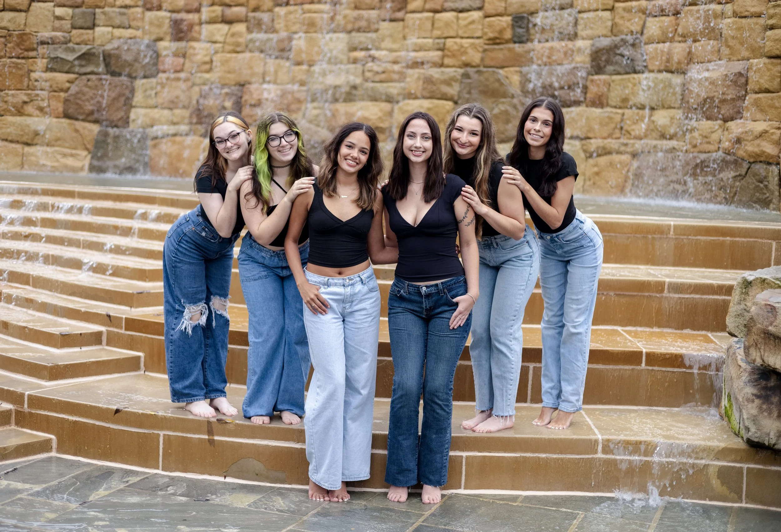 Group of six young women standing on a tiered stone fountain, smiling, with a brown brick wall behind them.