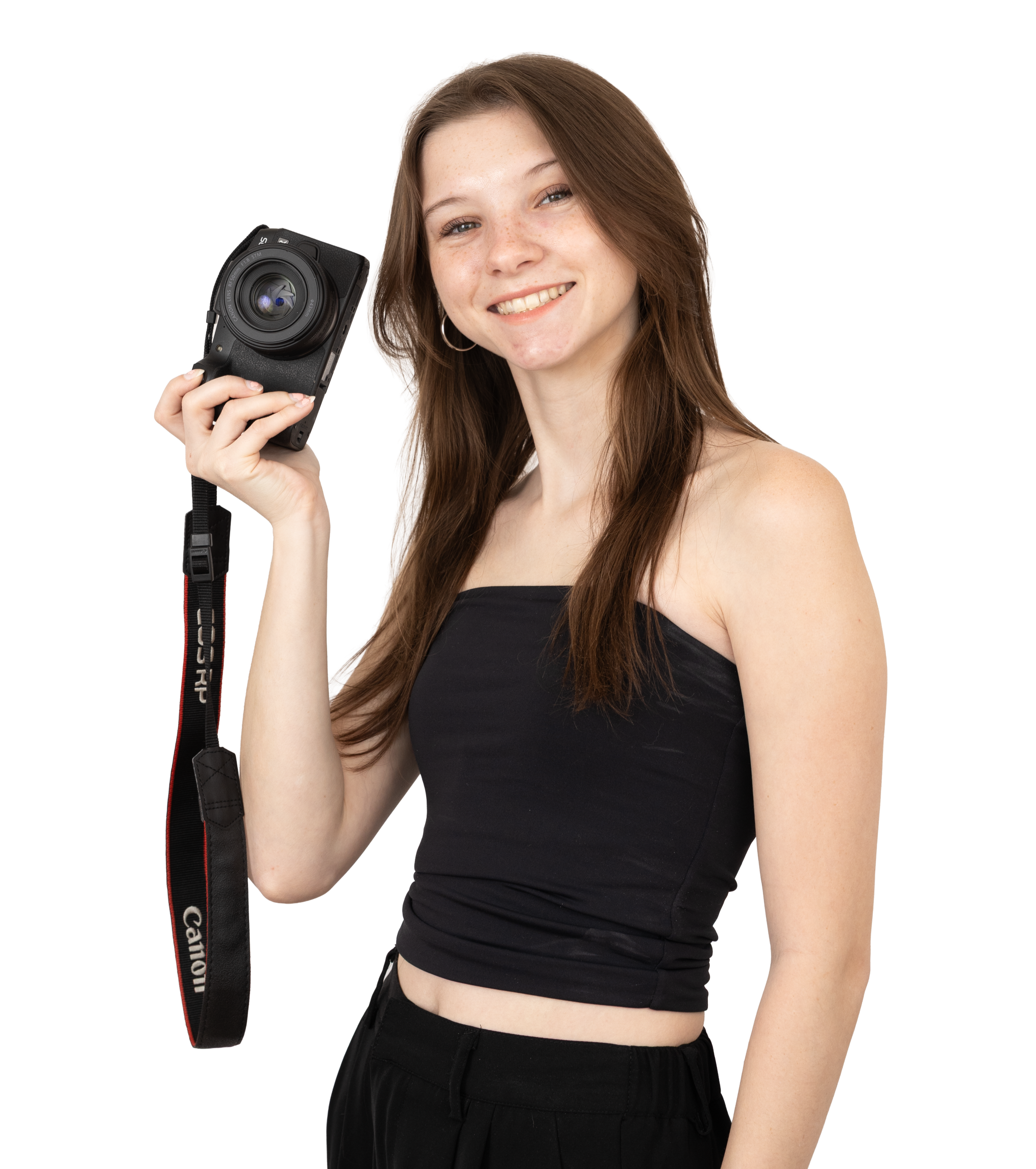 A young woman with long brown hair, wearing a black crop top and black pants, smiling and holding a Canon camera with an attached strap in her right hand.