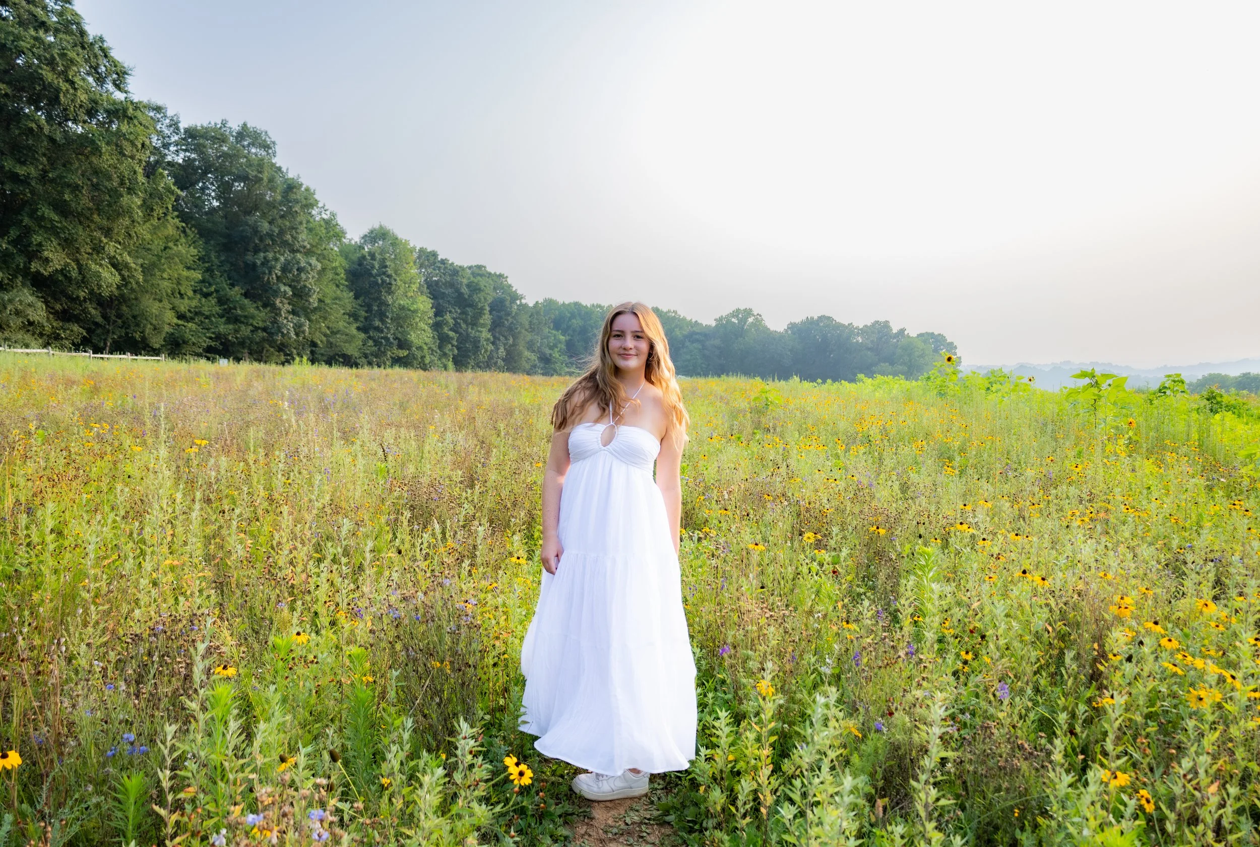 A young woman in a white dress standing in a wildflower field on a bright, sunny day.