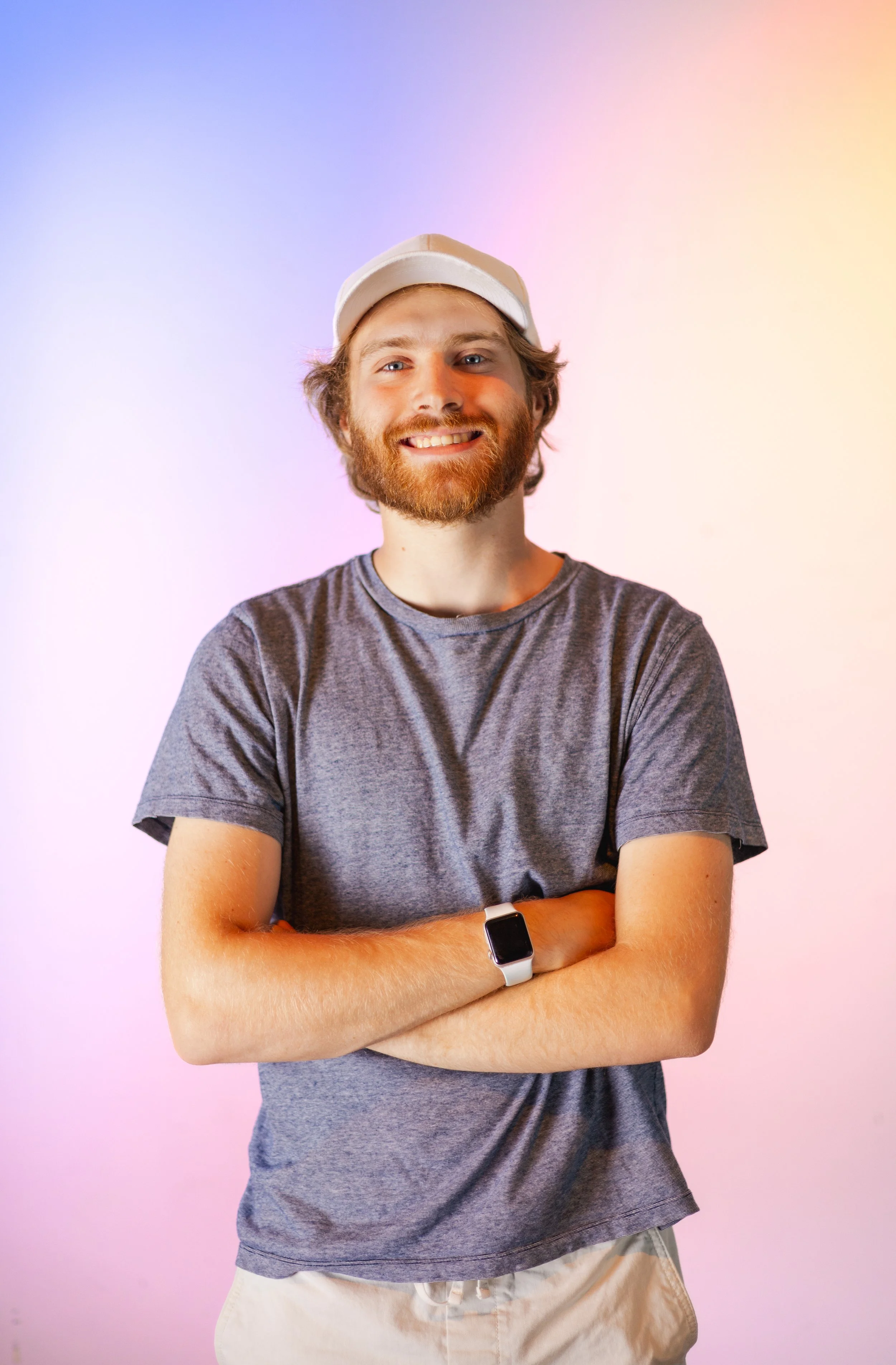 A young man with a beard and mustache is smiling, wearing a gray t-shirt, a white smartwatch, and a white cap. He has crossed arms and is standing against a pastel-colored background.