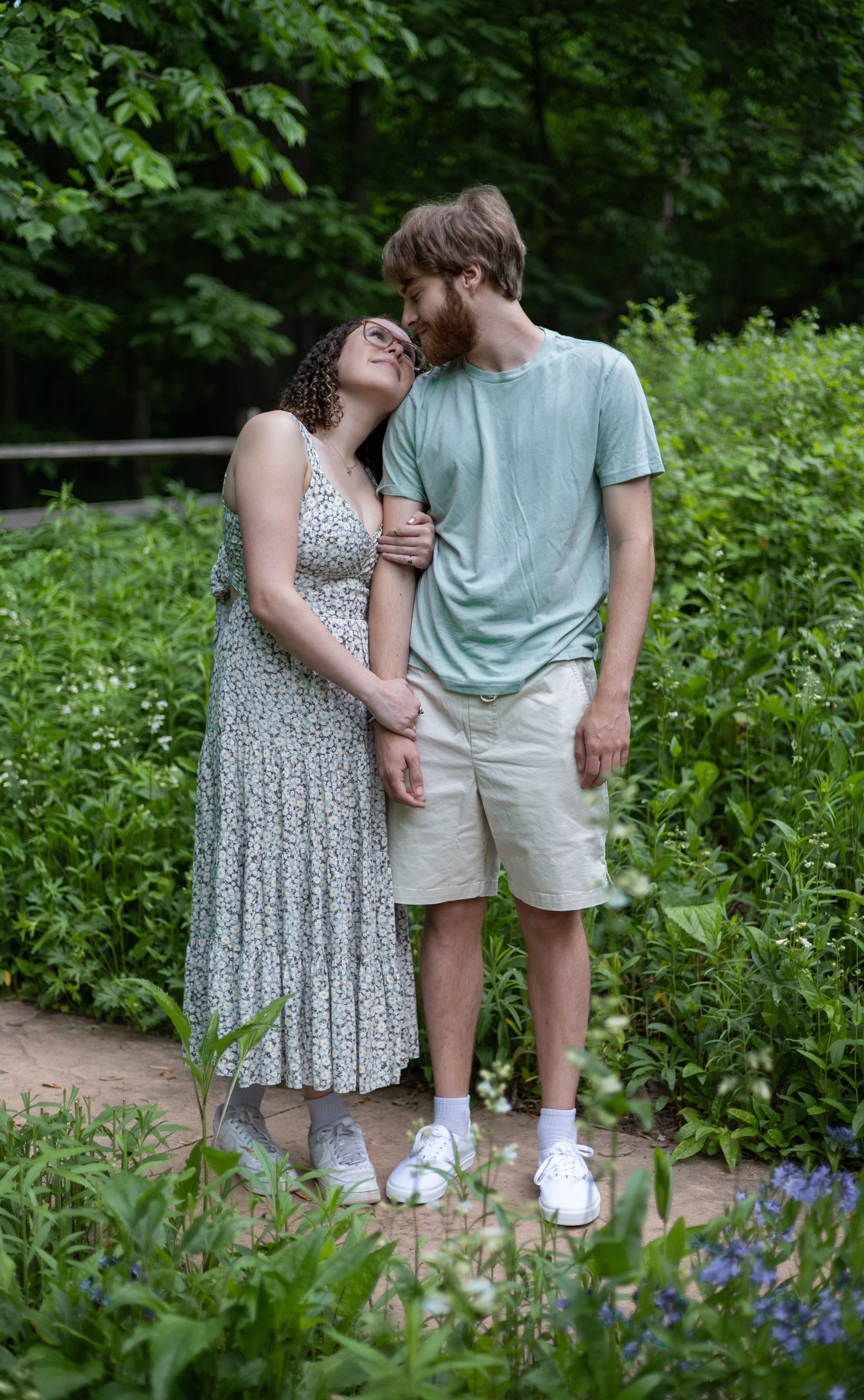 A young couple standing close together in a lush, green outdoor setting, with the woman affectionately leaning her head on the man's forehead, both smiling gently.