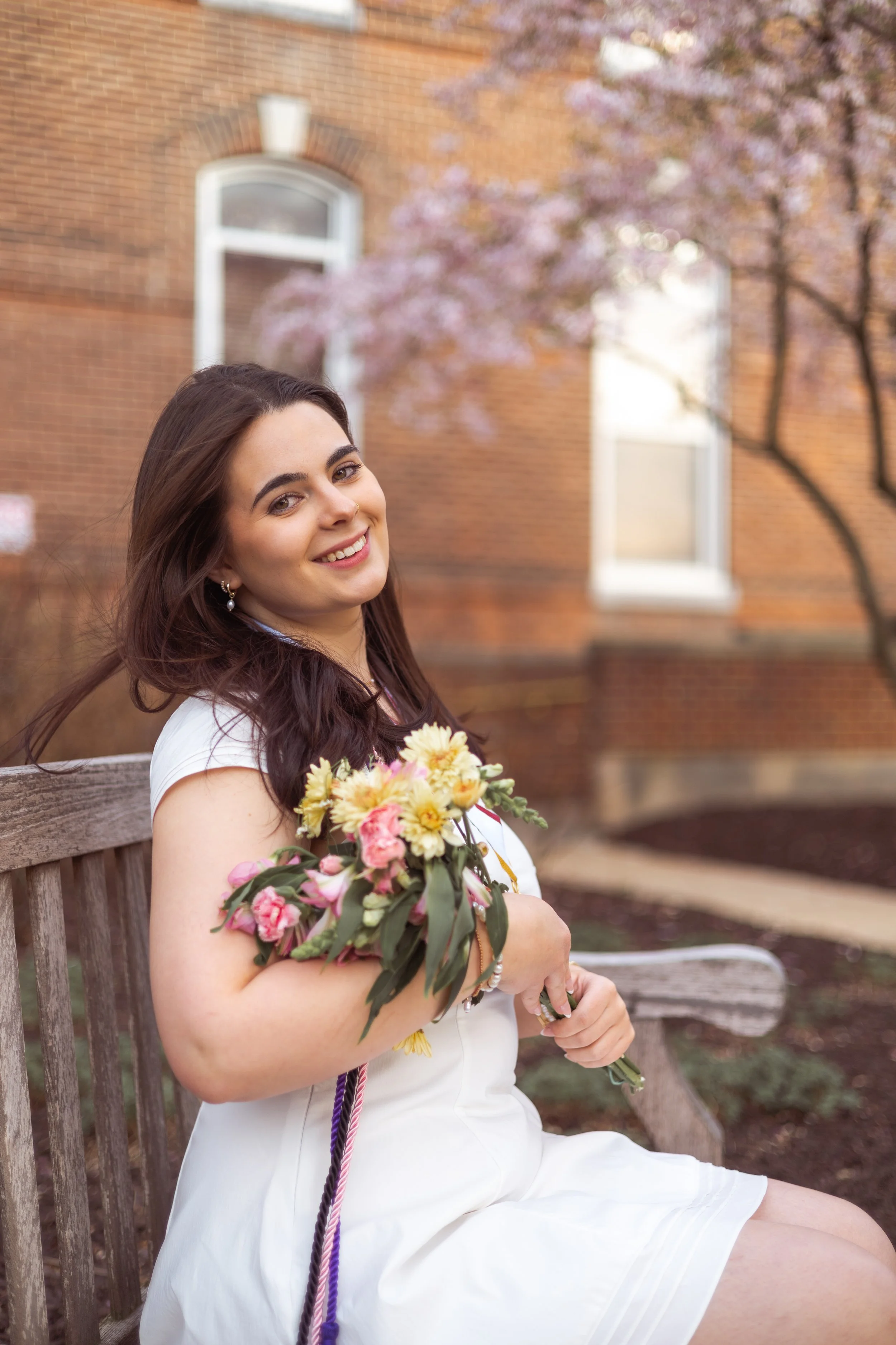 A woman with long dark hair sitting on a wooden bench outside, holding a bouquet of flowers, with pink flowering trees and a brick building in the background.