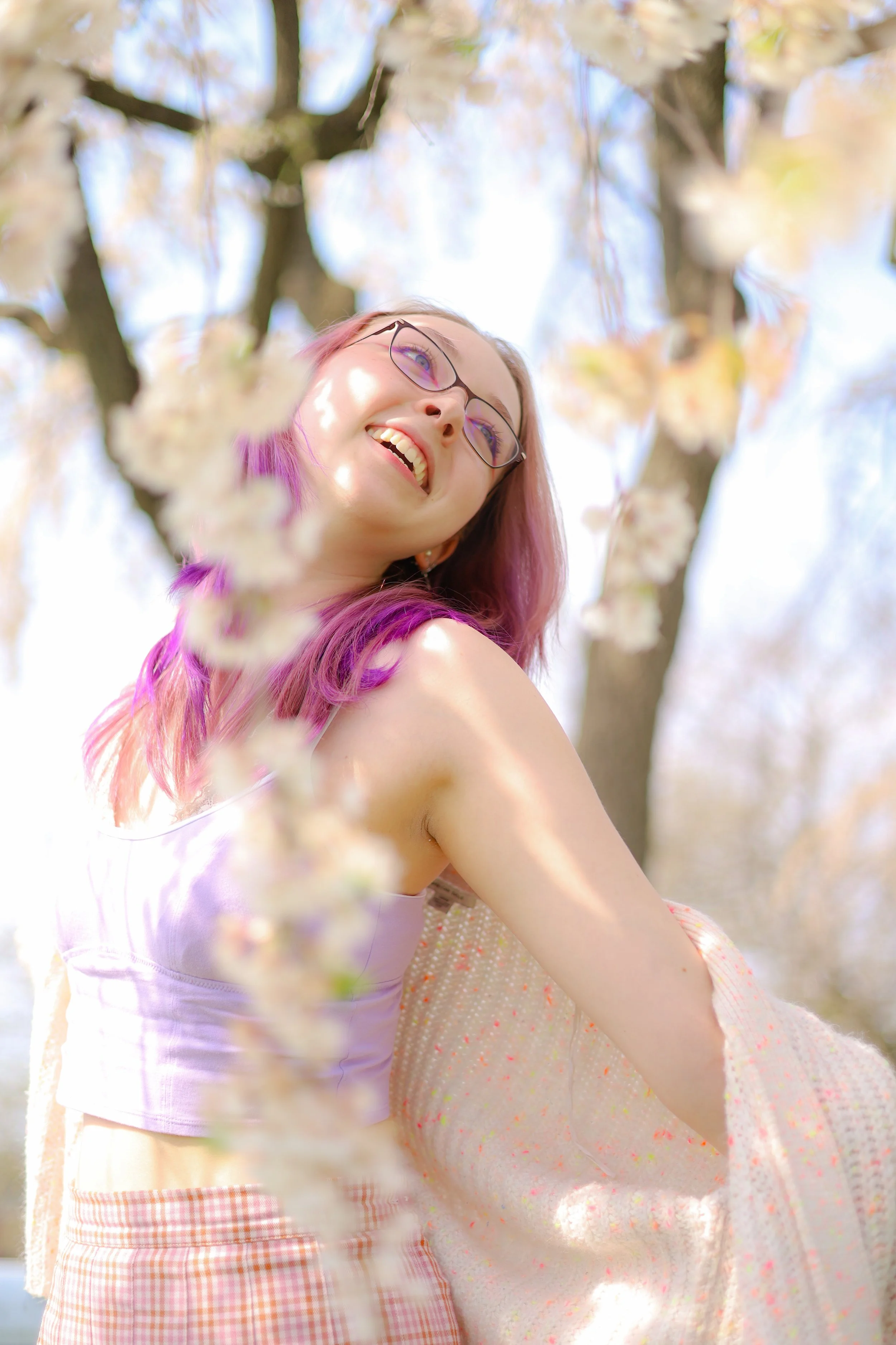 A young woman with pink and purple hair, wearing glasses and a light purple top, smiling and looking up, surrounded by blooming pink and white flowers on a tree with a bright, sunny sky in the background.