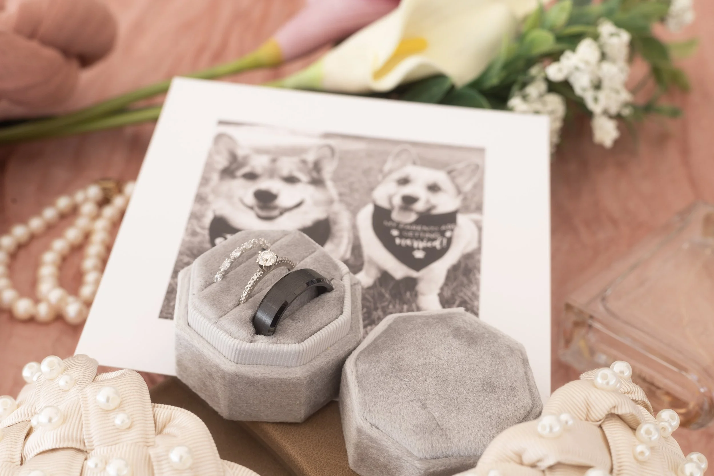A photograph of wedding rings and jewelry on a pink surface with flowers in the background. There are two rings in a gray velvet box, one with diamonds and the other a black band, along with decorative items such as pearl-adorned shoes, a pearl neckl