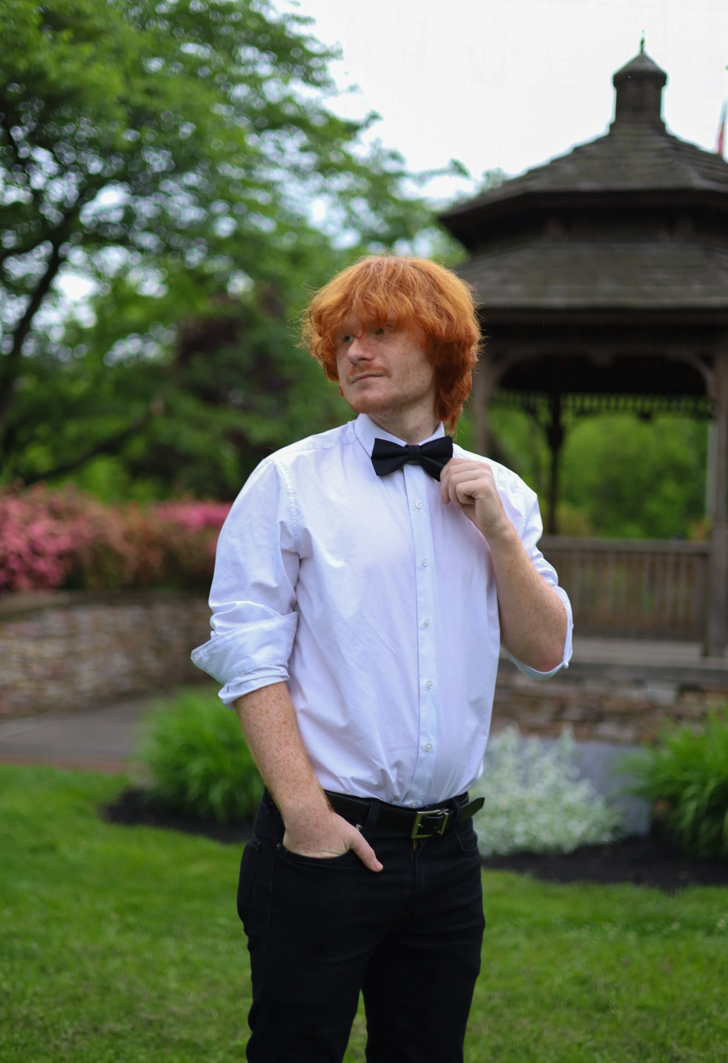 A man with curly red hair and freckles, wearing a white dress shirt with rolled-up sleeves, black pants, and a black bow tie, stands outdoors in front of a wooden gazebo. He has one hand in his pocket and the other adjusting his bow tie, with a thoug