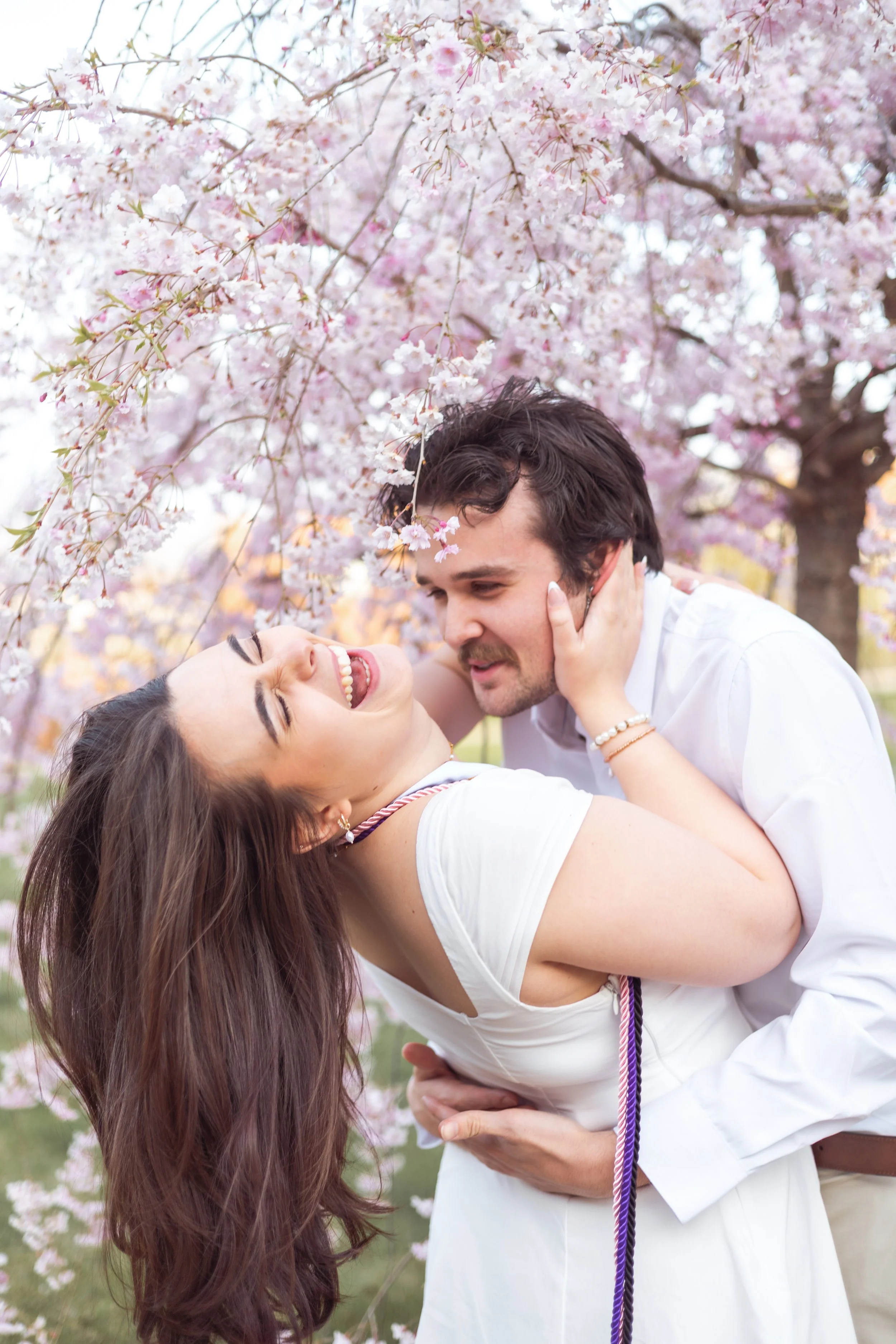 A couple sharing a joyful moment outdoors under cherry blossom trees in bloom, with the woman laughing and leaning back, and the man holding her.
