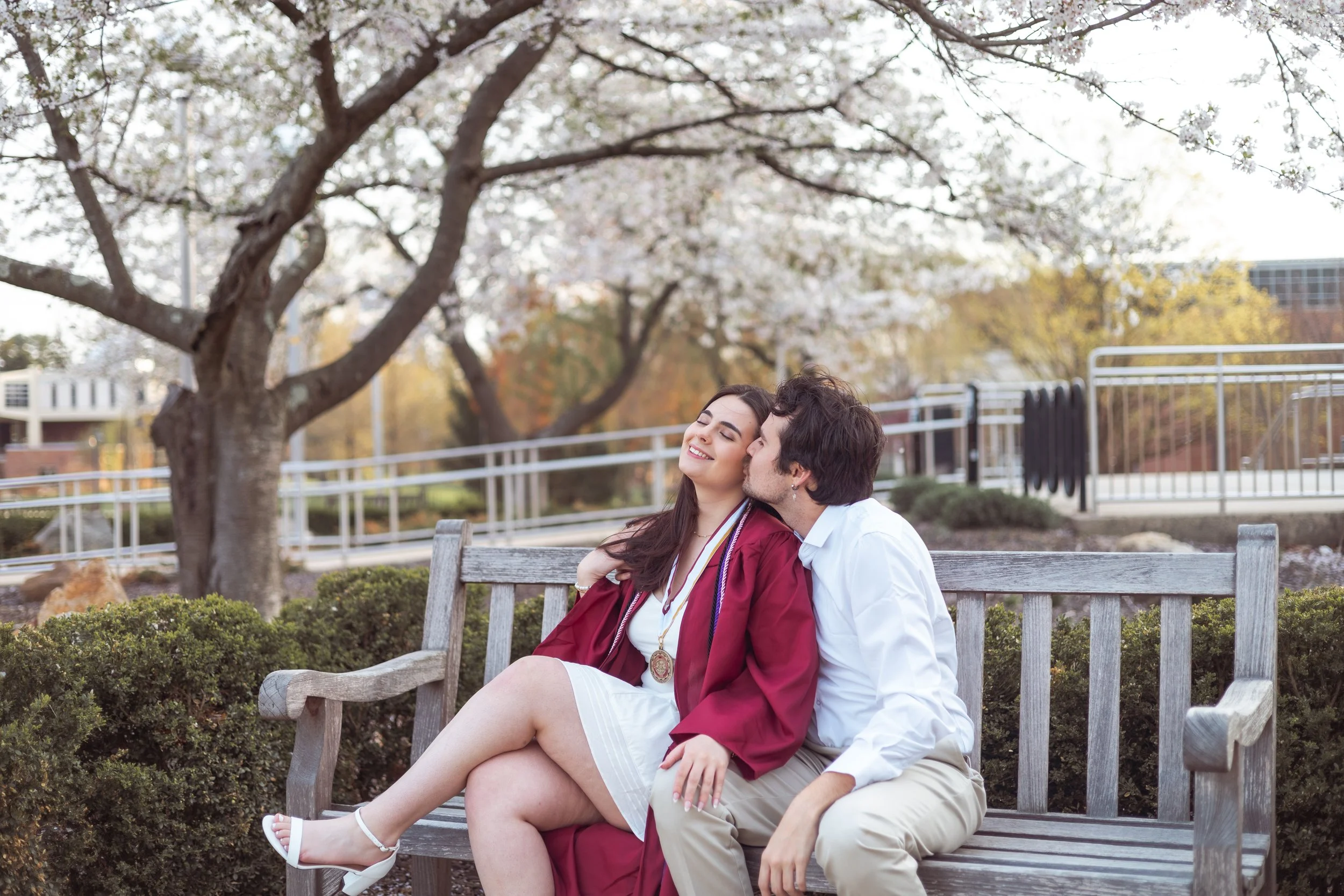 A young woman in a white dress and maroon graduation gown sits on a park bench while a young man kisses her cheek, surrounded by blooming cherry blossom trees.