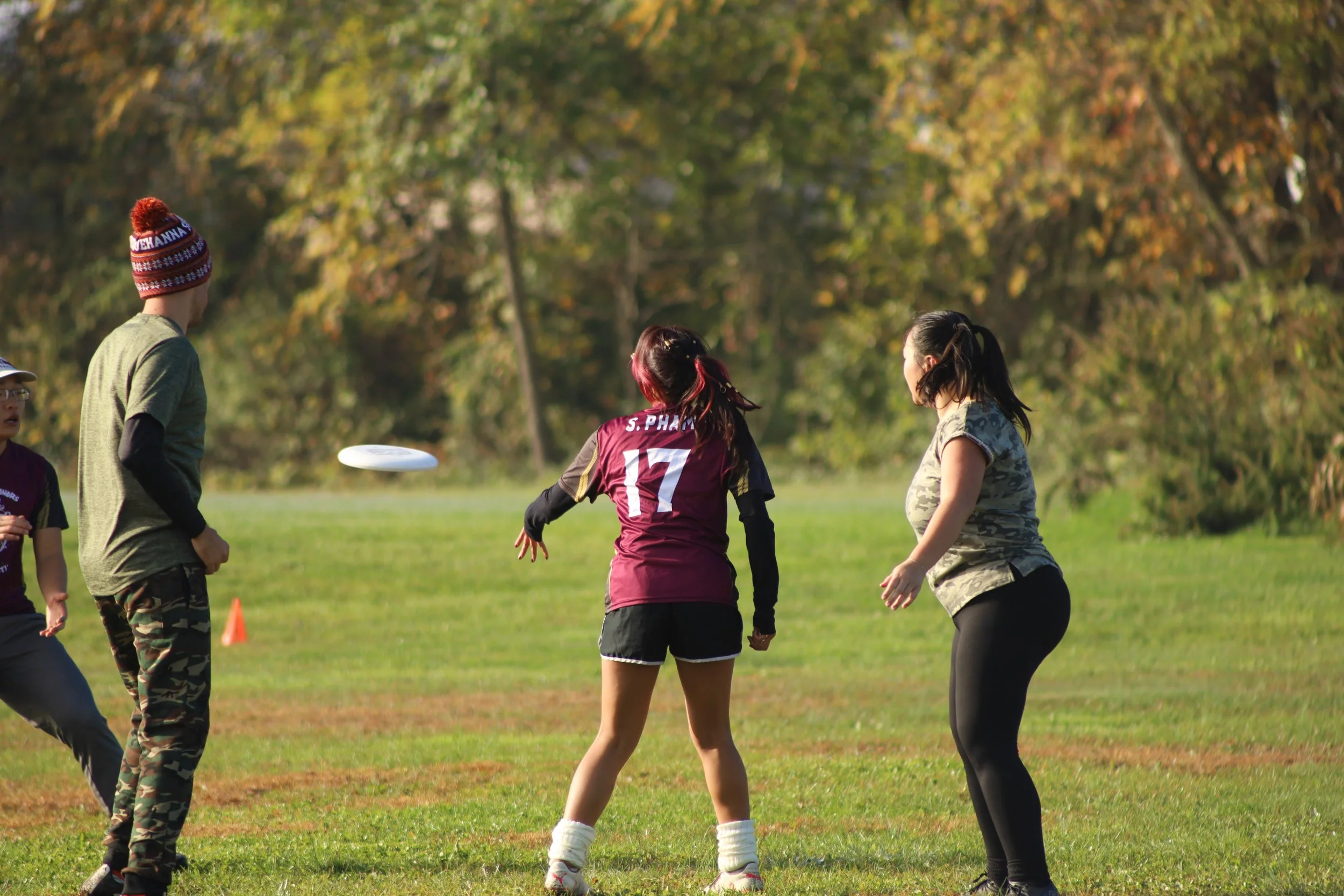 Four people playing frisbee on a grassy field with trees in the background during fall.