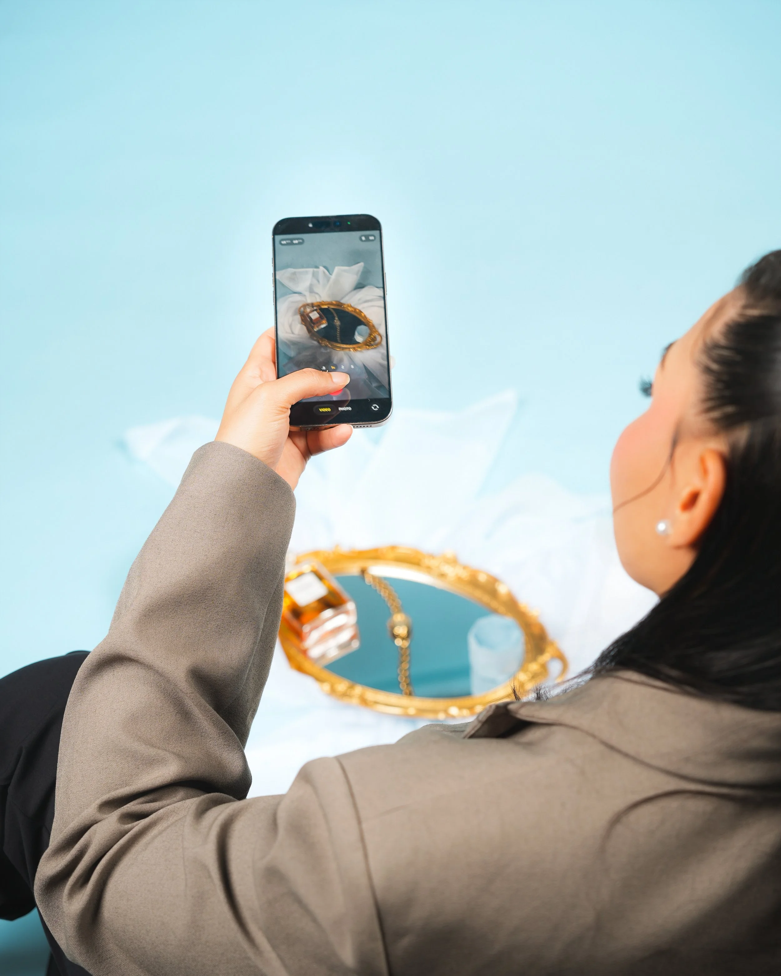 A woman is taking a photo of a gold-framed mirror with her smartphone, showing a bracelet and a chain inside the mirror.