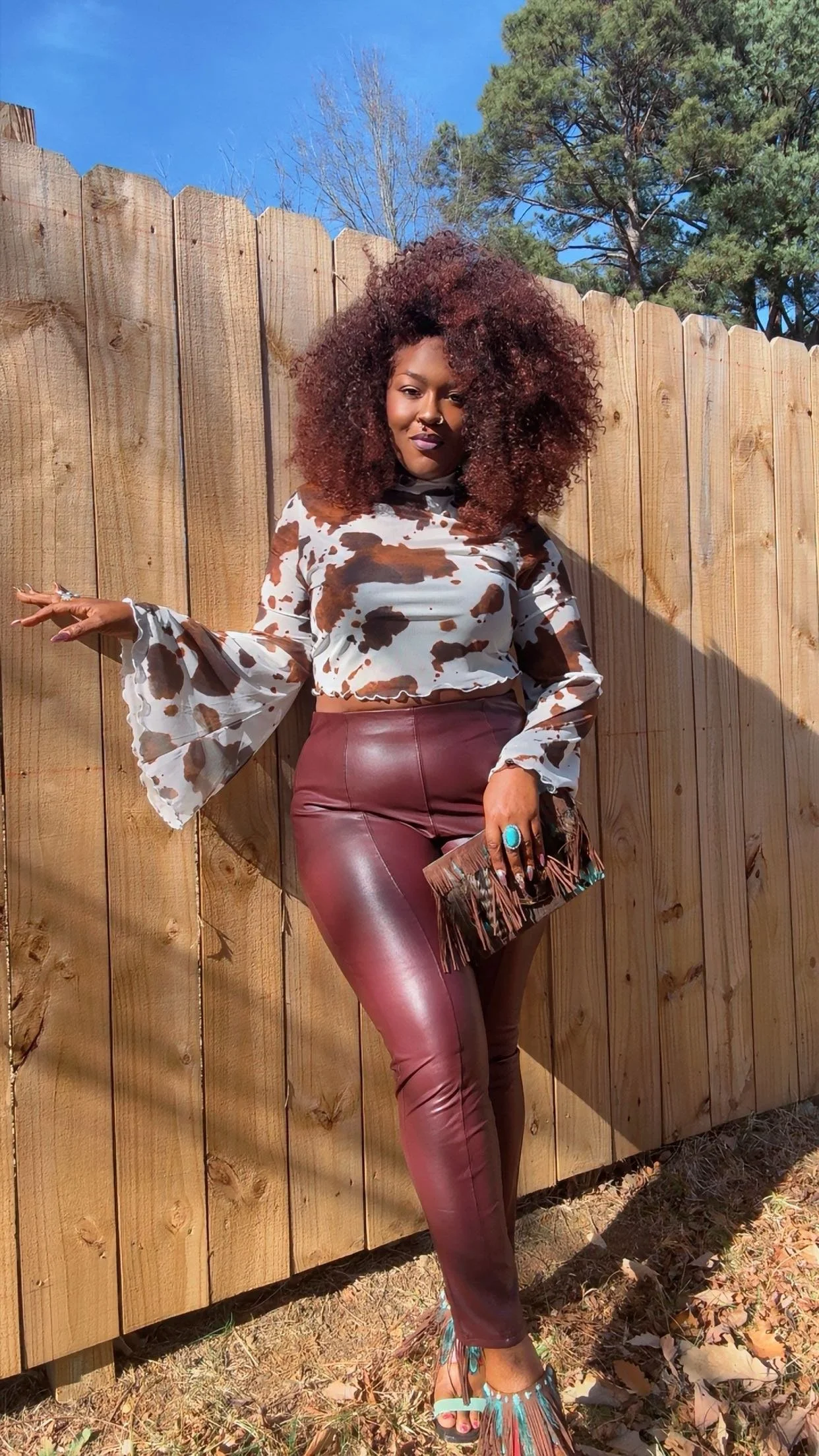 A woman with voluminous curly hair standing outdoors against a wooden fence, wearing a cow-print silk blouse, maroon leather pants, colorful fringed heels, and holding a small fringed clutch.