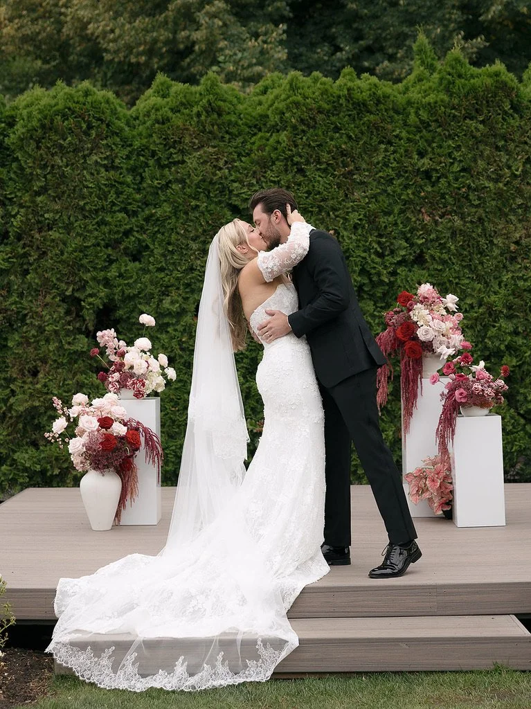 Bride and groom kissing during their garden wedding at Columbia Collective, surrounded by romantic pink, burgundy, and blush wedding florals displayed on modern white pedestals. Floral design by Butterfly’s Rest. Photography by Mercedes Lou Photograp