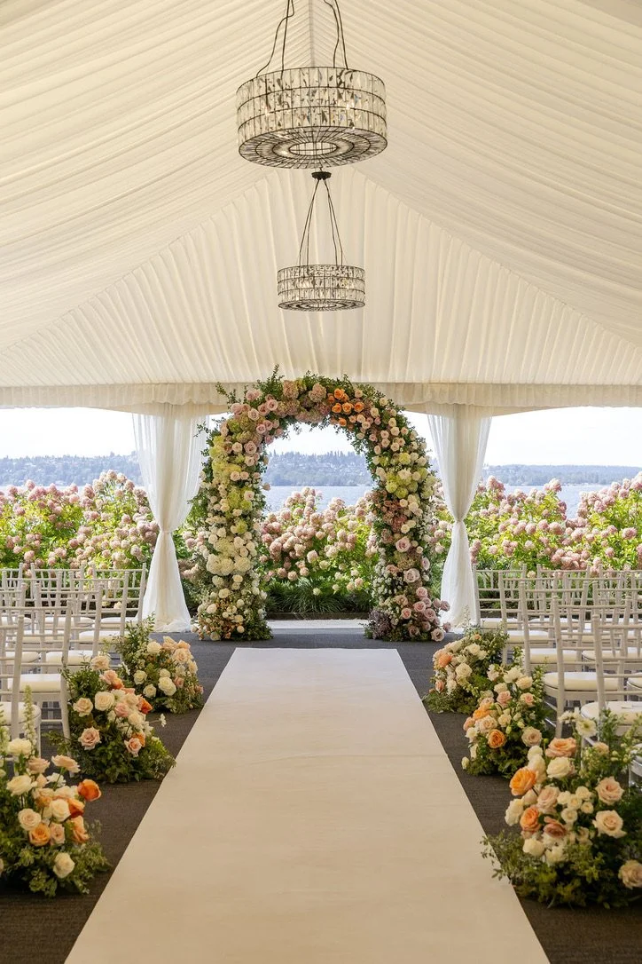 Romantic wedding ceremony featuring a lush floral arch and garden-inspired aisle arrangements in blush, peach, cream, and green overlooking the water at The Woodmark Hotel. Planning & floral design by Luth Weddings & Events. Floral lead by Butterfly’