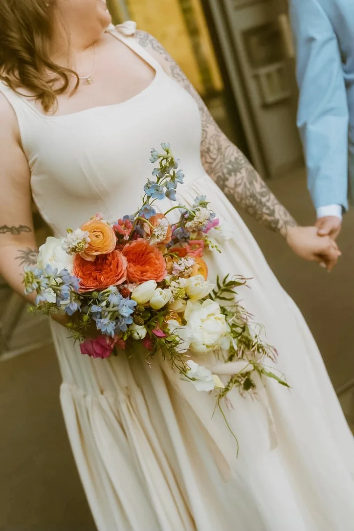 Bride holding a colorful, garden-inspired cascading bouquet featuring orange, peach, blue, pink, and white seasonal blooms for the elopement. Floral design by Butterfly’s Rest.