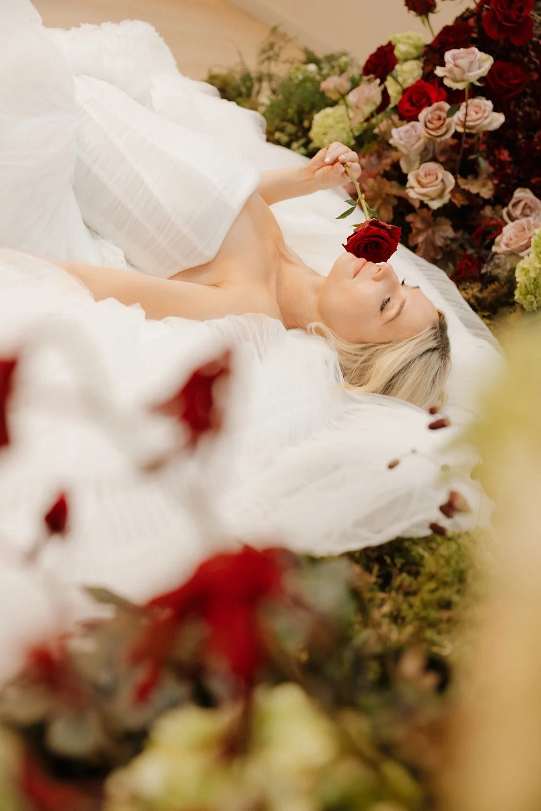Bride holding a red rose while resting among lush wedding florals and soft layers of tulle.