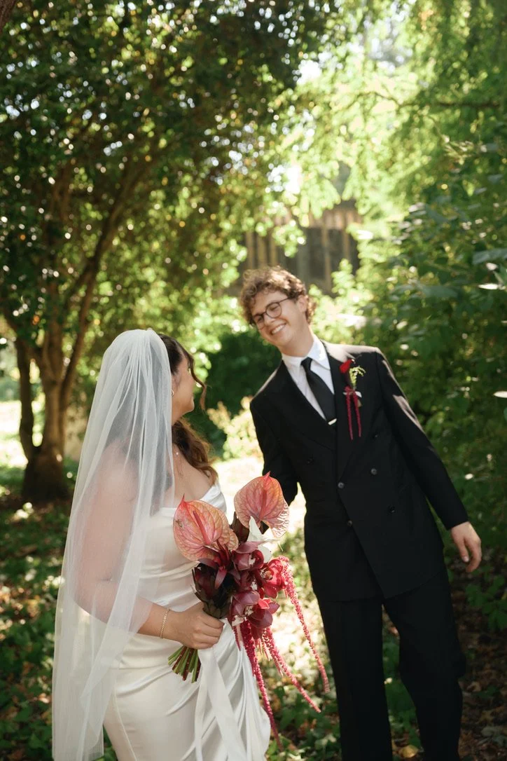 Bride holding a sculptural bridal bouquet featuring red and blush anthuriums, cascading burgundy amaranthus, and textural seasonal foliage. Floral design by Butterfly’s Rest. Photography by Elita Coralee.