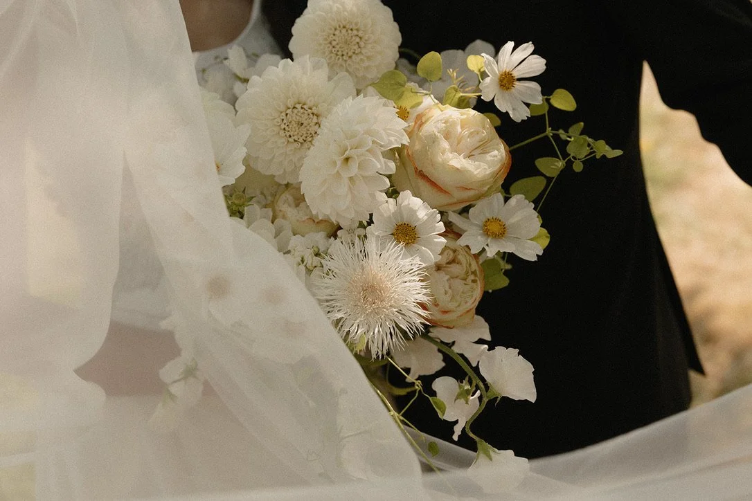 Close-up of an elegant white bridal bouquet featuring dahlias, garden roses, cosmos, and textural seasonal blooms, softly framed by the bride’s veil. Floral design by Butterfly’s Rest. Photography by Tori Osteraa Photography.
