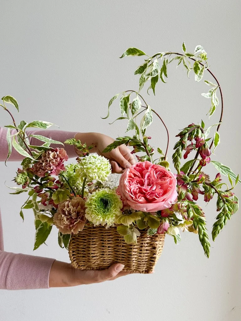 Wicker basket arrangement featuring seasonal flowers and lush greenery in shades of pink, white, and green.