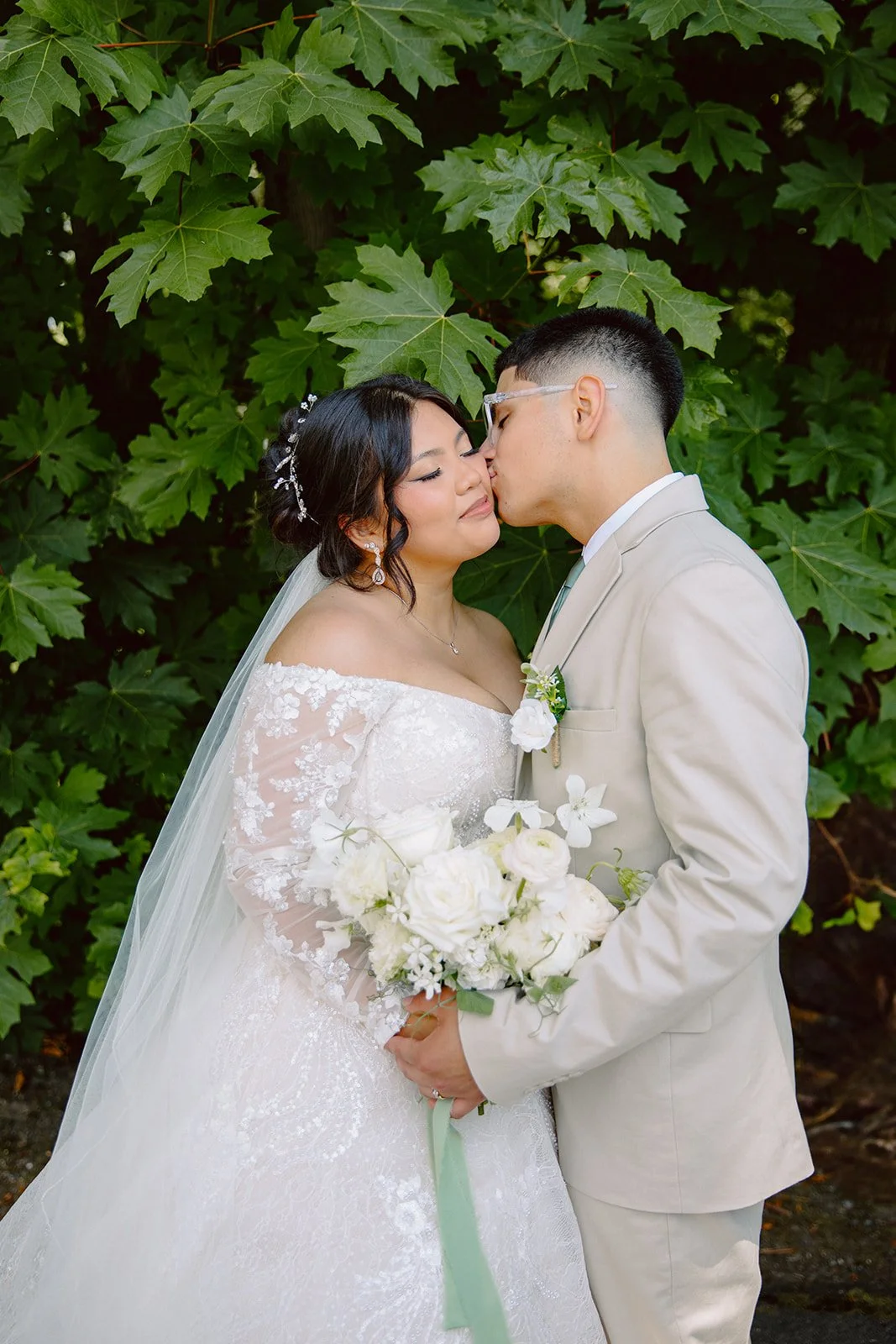 Bride and groom sharing a kiss surrounded by greenery, with the bride holding an elegant white bridal bouquet.