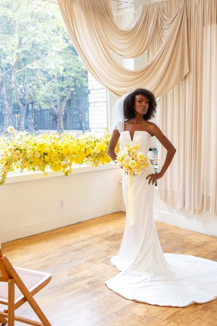 Bride holding a butter yellow bridal bouquet beside a vibrant yellow hanging floral installation in a light-filled wedding setting at 210 Seattle. Floral design by Butterfly’s Rest. Photography by Daymark Studios.