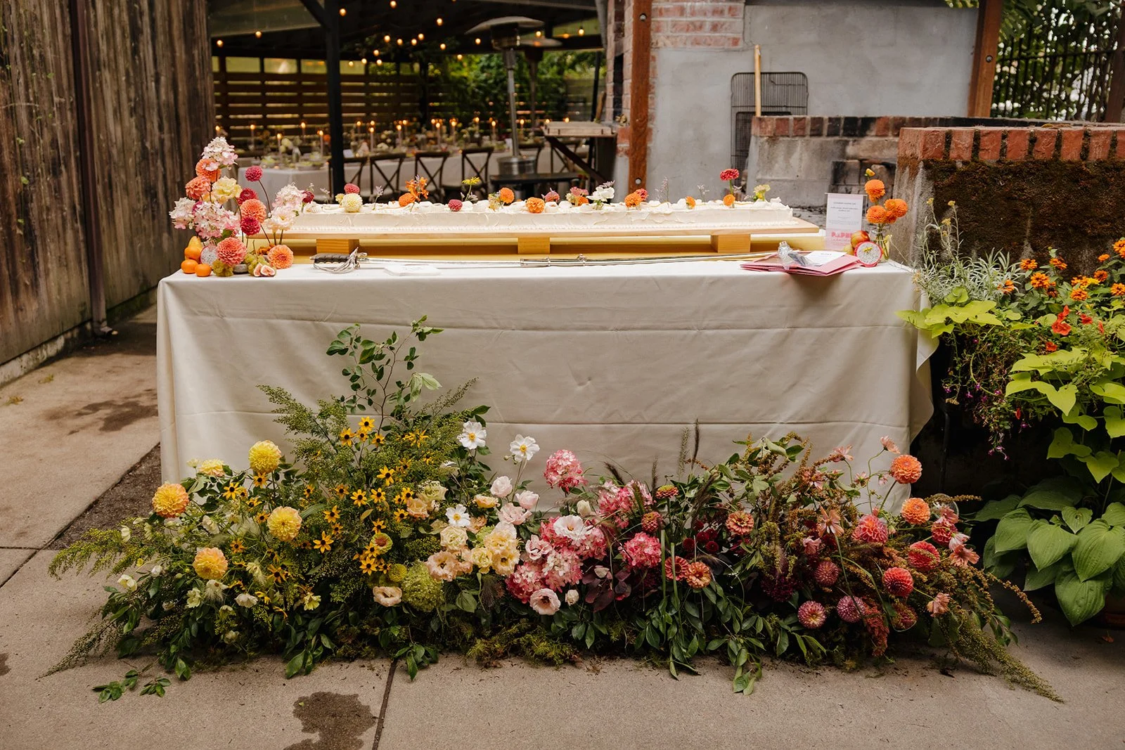 Wedding cake table featuring abundant seasonal floral ground installations in vibrant late-summer tones.