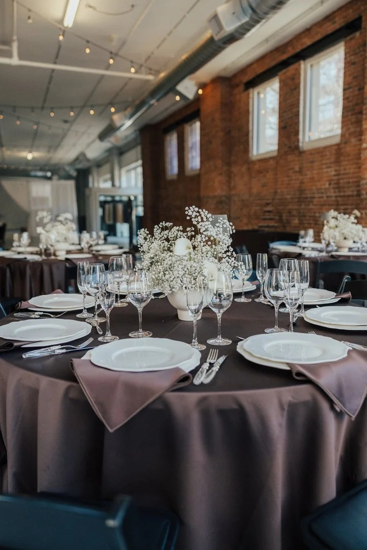 Round reception table featuring a cloud-like white floral centerpiece, elegant place settings, and a dark linen tablecloth.