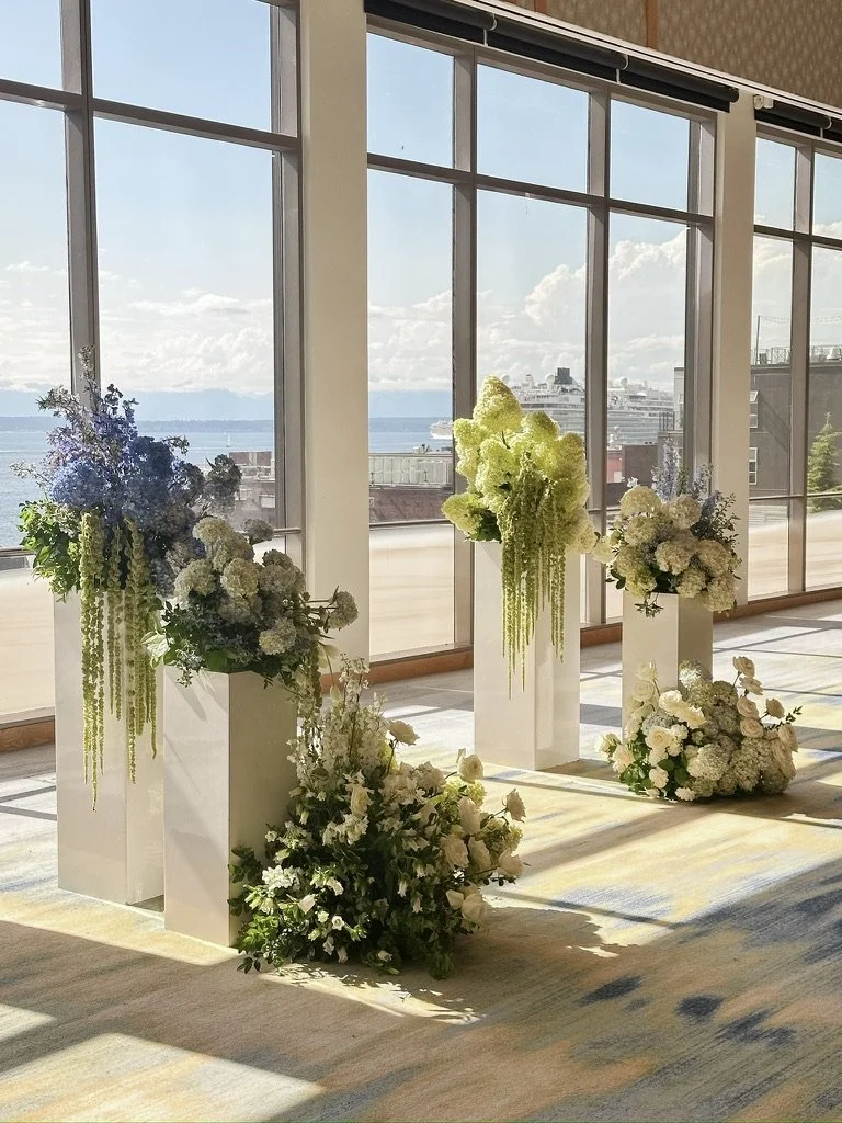 Sculptural wedding floral installations displayed on white pedestals with cascading blue, white, and green seasonal blooms overlooking the Seattle waterfront at Four Seasons Hotel Seattle. Planning & floral design by Luth Weddings & Events. Floral le